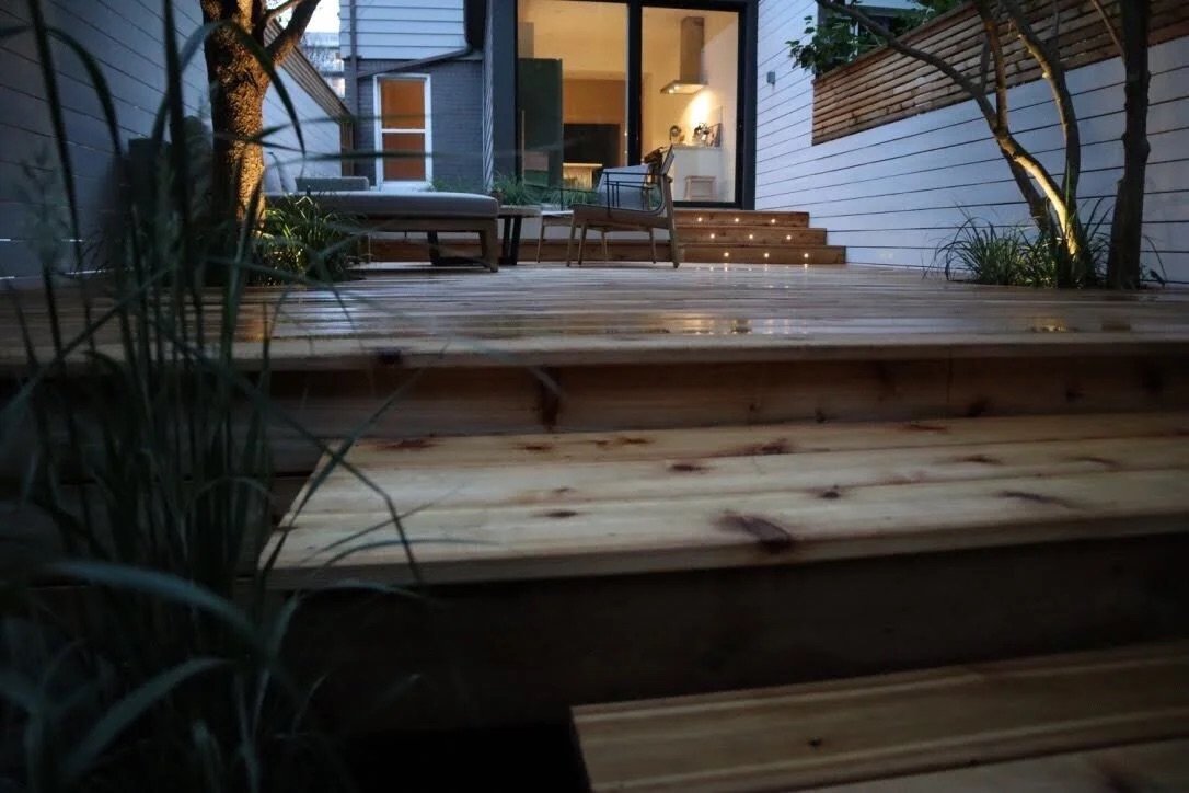 View of a wooden deck with stairs leading up to a house with large glass doors, outdoor seating, and interior lighting.