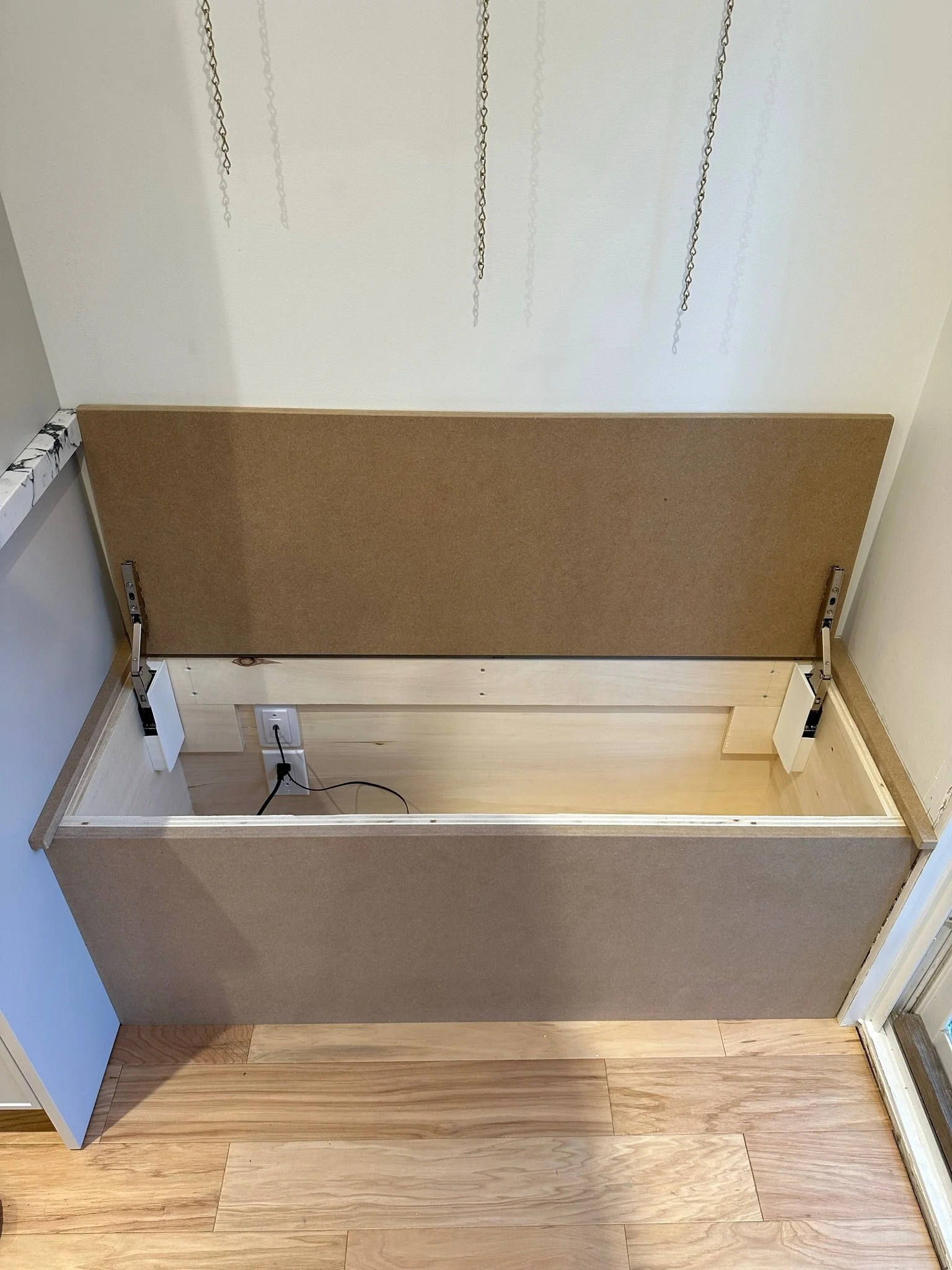 An empty hatchback bed with a corkboard attached to the back, located in a room with wooden flooring, a power outlet, and cables connected to devices.