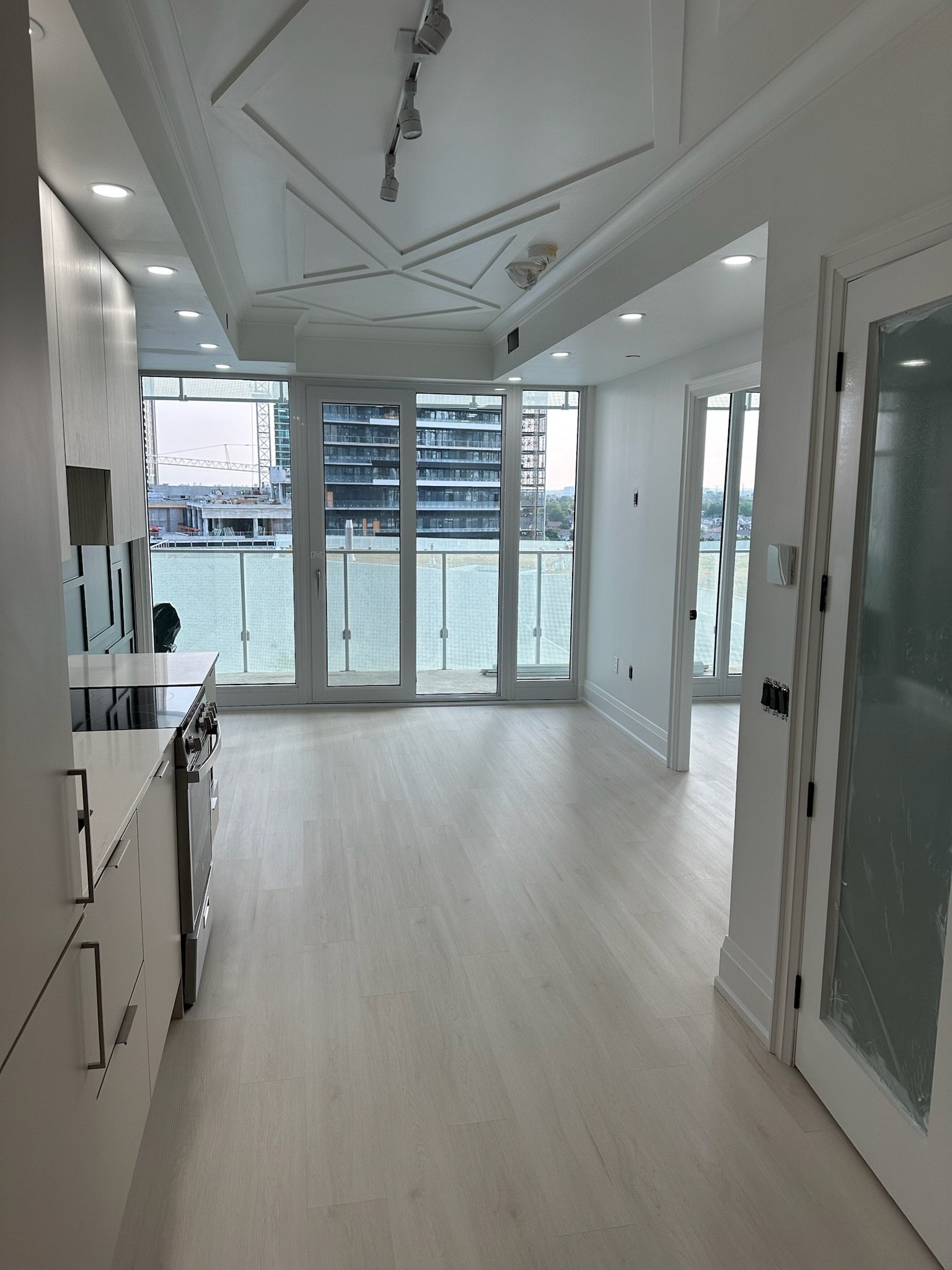 Modern kitchen with white cabinets, light wood floors, large glass doors leading to balcony, and city view in background. There is detailed carpentry and trim work on the ceiling.
