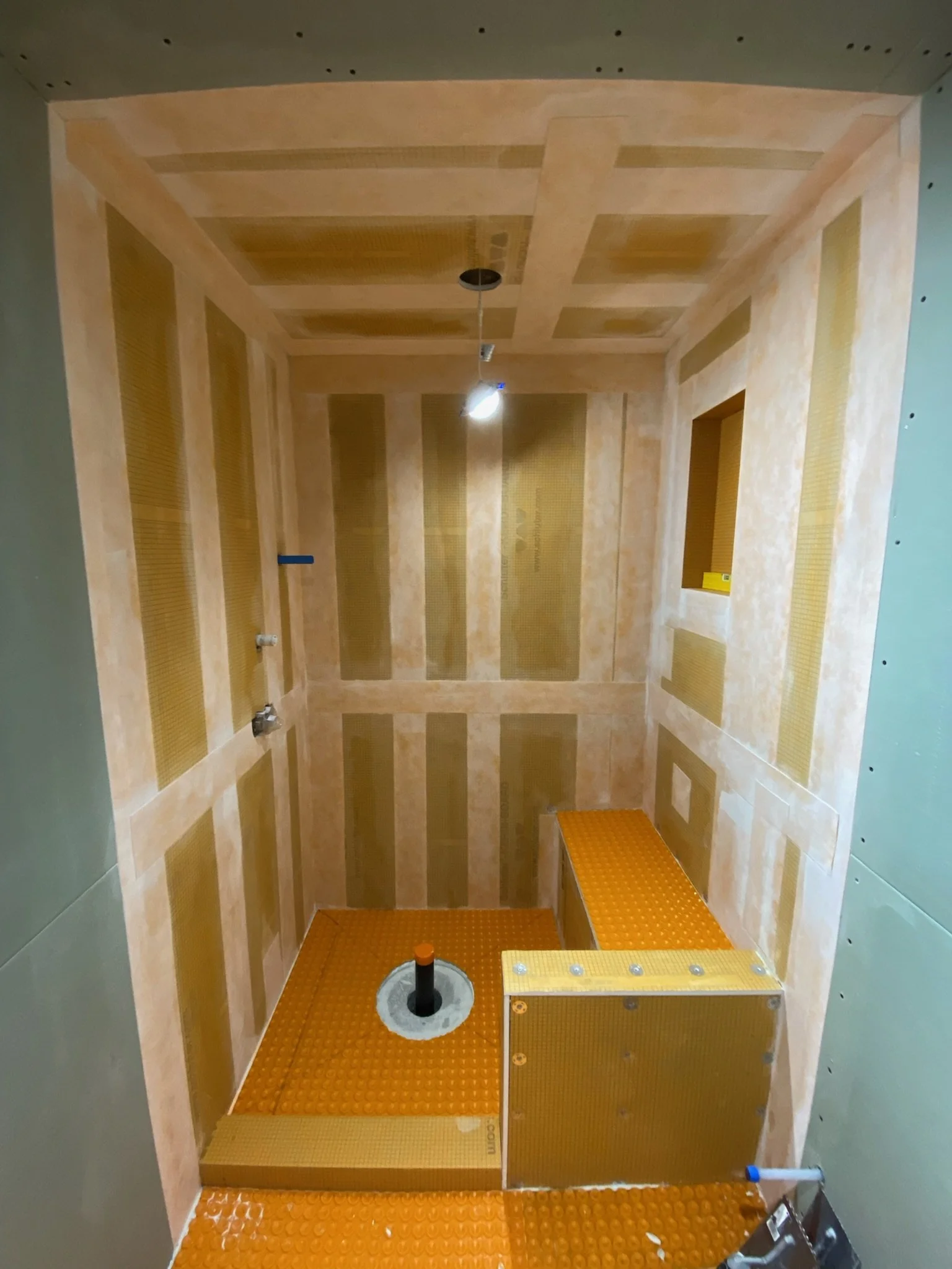 The image shows an unfinished shower with a wood frame, waterproofing, and a partially installed plumbing pipe in the center. The walls are not yet tiled, but the structure for tiling is meticulously in place.