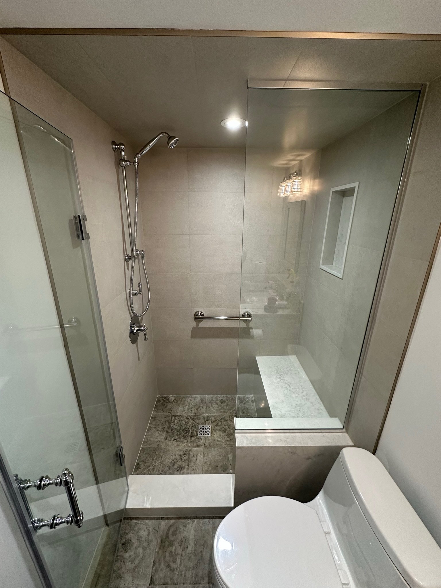 A modern bathroom with a walk-in shower, chrome fixtures, beige tiled walls, a built-in shelf, a toilet, a mirror, and a ceiling light.