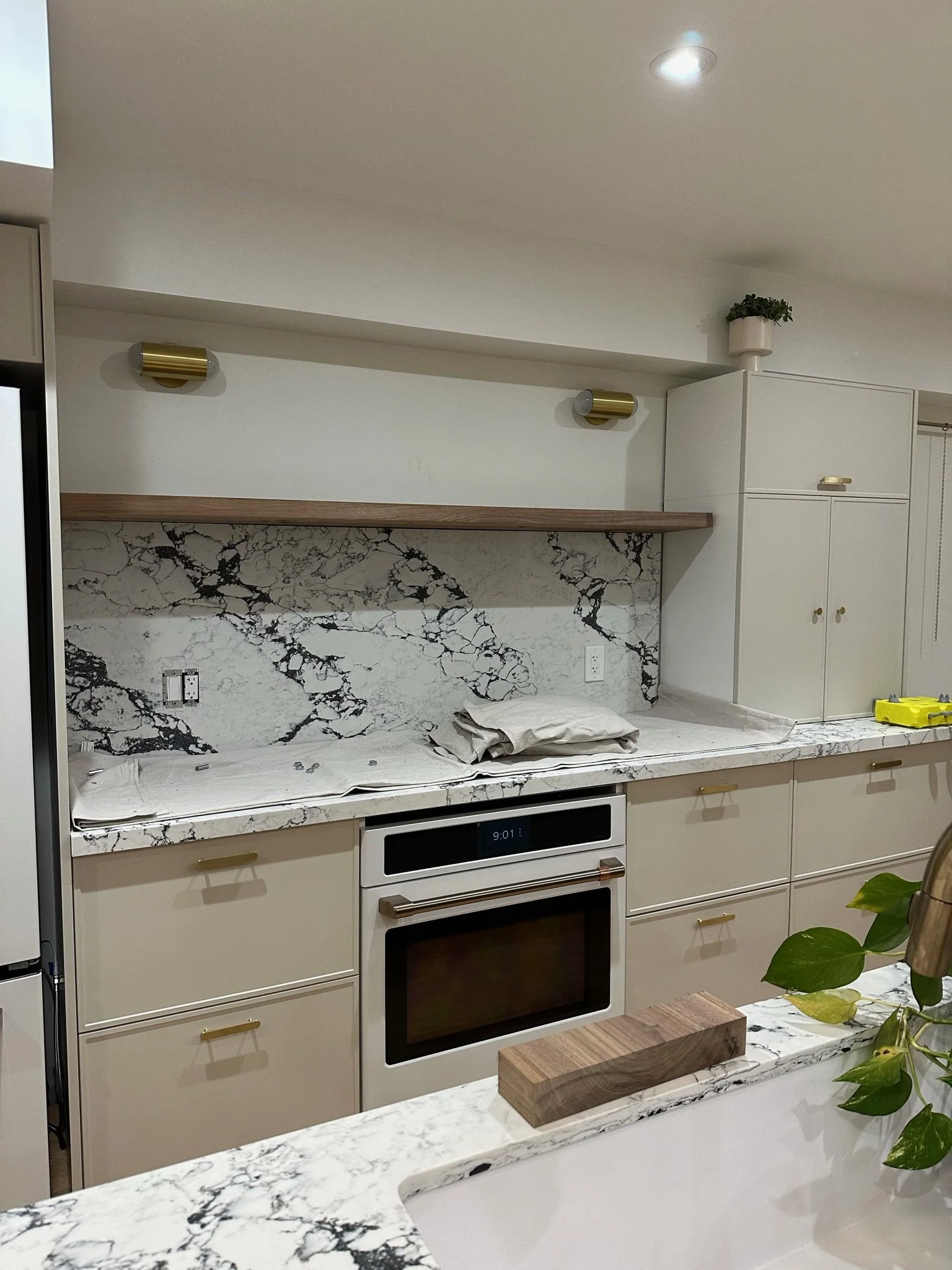 Modern kitchen with white cabinets, marble countertops, and a marble backsplash. There are two gold wall sconces above the countertop, a potted plant on top of a cabinet, and a yellow tool on the counter. An oven with a digital clock showing 9:01 is built into the cabinetry.