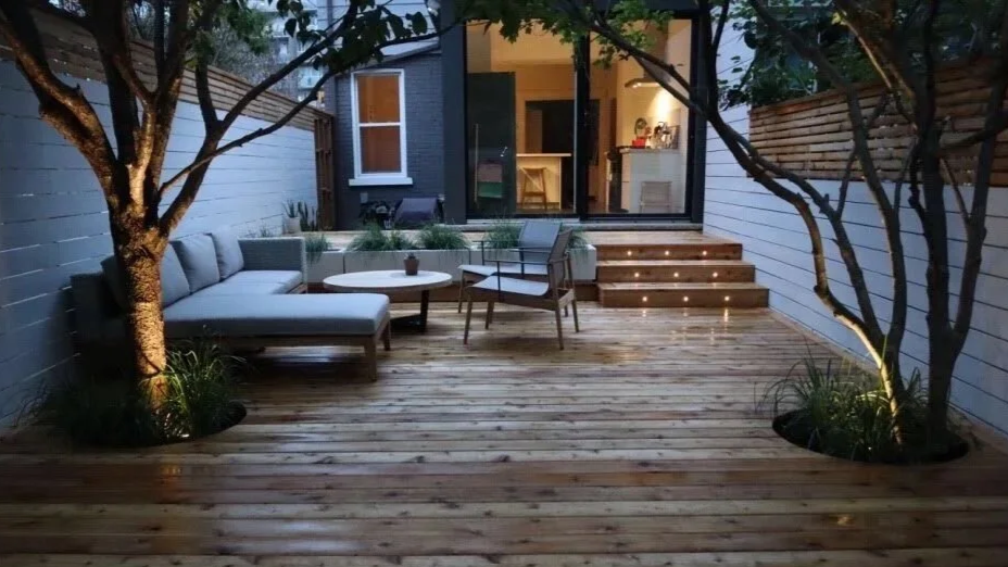A cozy outdoor patio with wooden decking, two small trees with lighting, a seating area with cushions, a round table, and a sliding door leading inside a house.