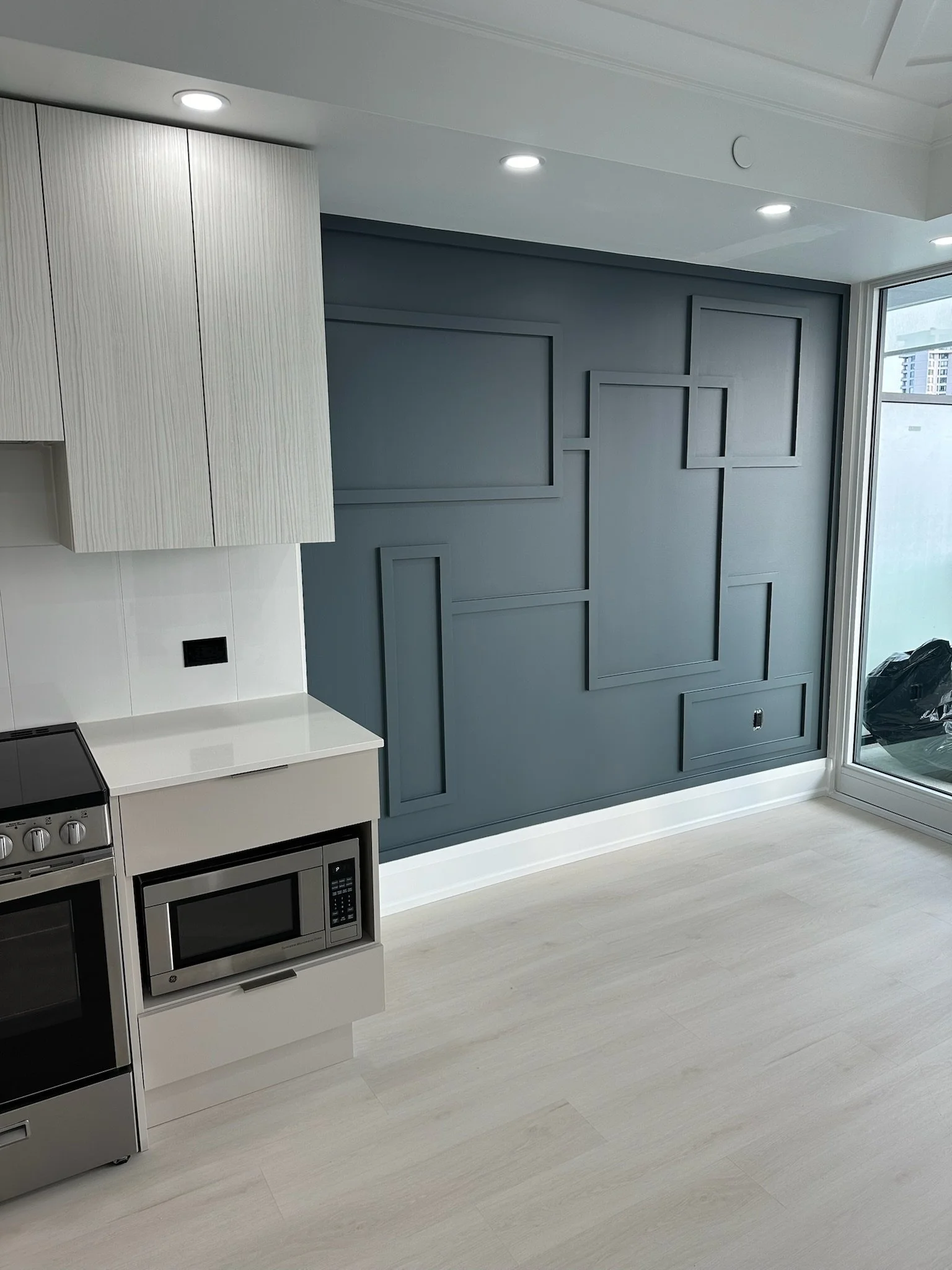 Modern kitchen with light-colored flooring, a white counter with a black stove and microwave, and a blue accent wall with decorative framing. There is a sliding glass door on the right letting in natural light.