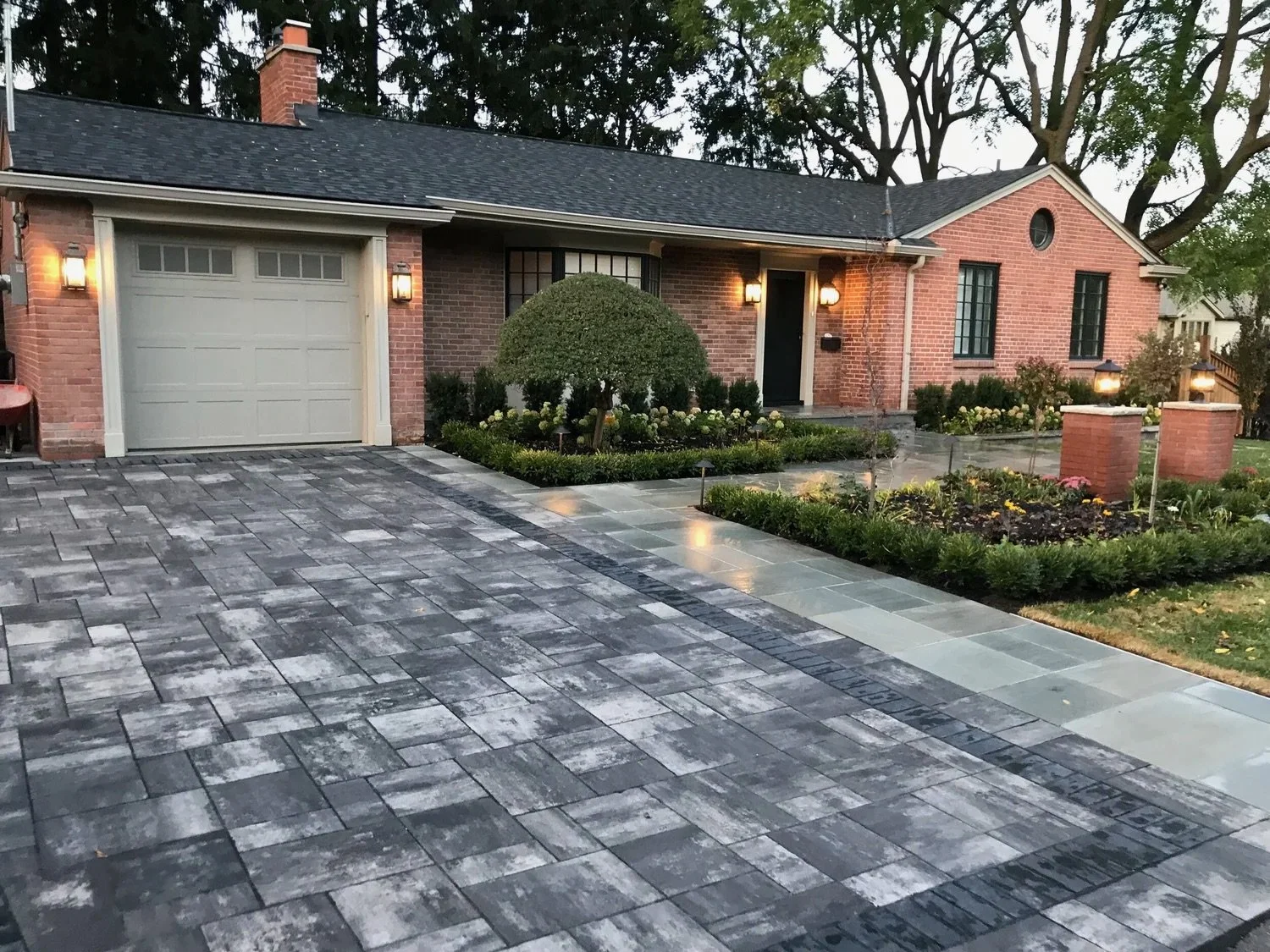 Red brick house with a dark shingle roof, multiple windows, a white garage door, and a beautifully landscaped front yard with a rounded bush, flowers, and a paved driveway. The stones on the driveway are incredibly neat with stunning lighting.