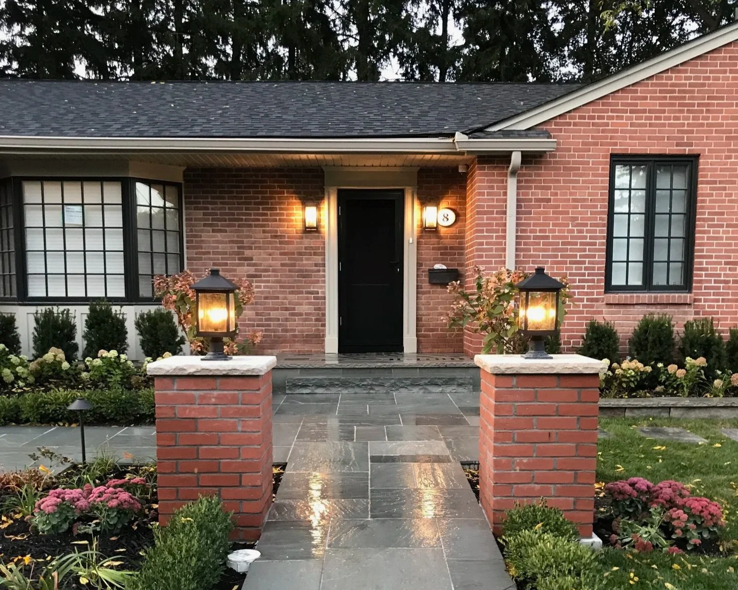 Front entrance of a brick house with a black door, two outdoor lanterns, and a small garden with colorful flowers, paved walkway, and brick pillars with additional lanterns.