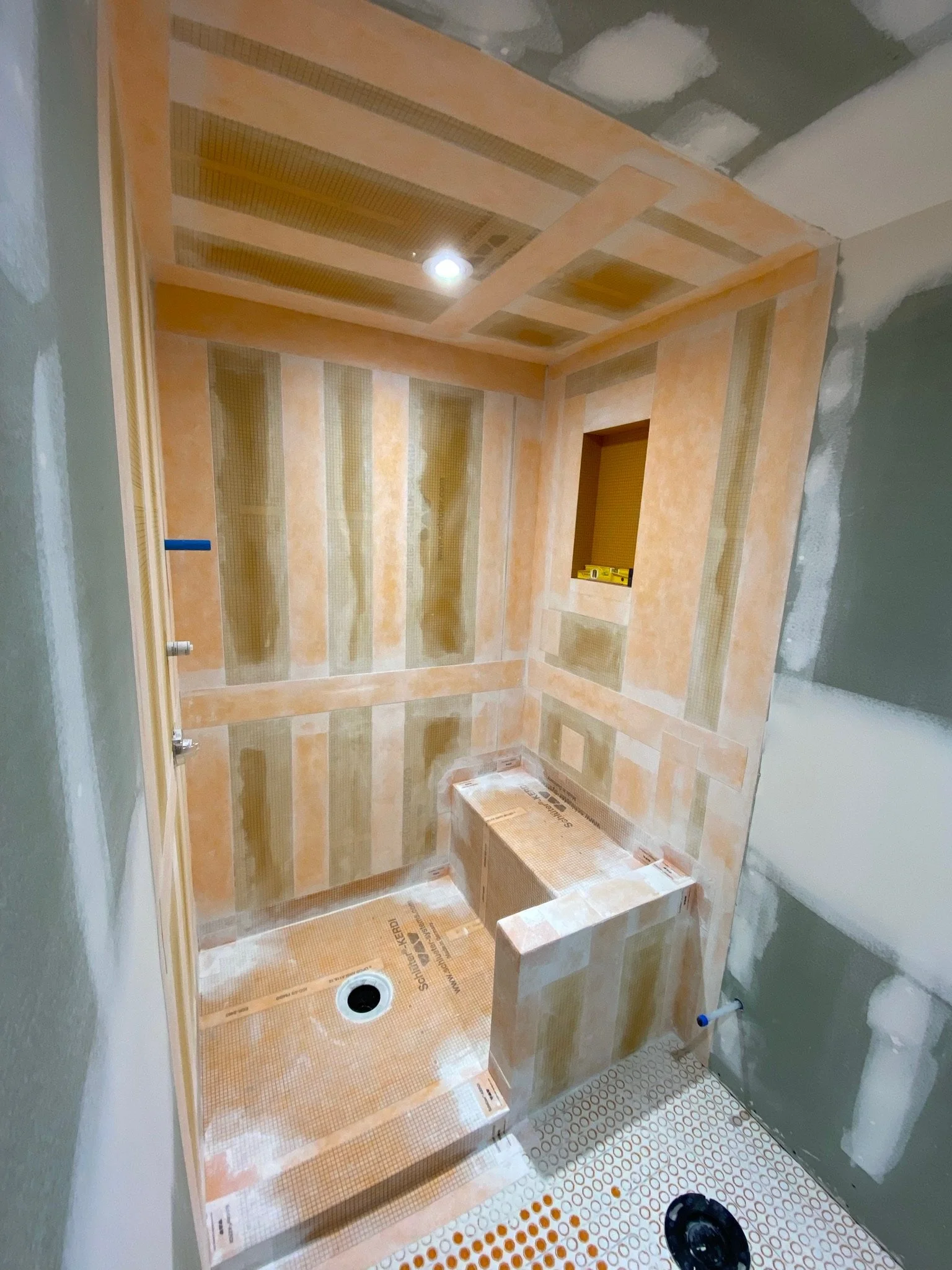 Bathroom under construction with new tile installation, including a shower area with a perfectly tiled wall and a floor with circular tile pattern. A black drain cover is visible on the floor, and a blue pipe protrudes from the wall.