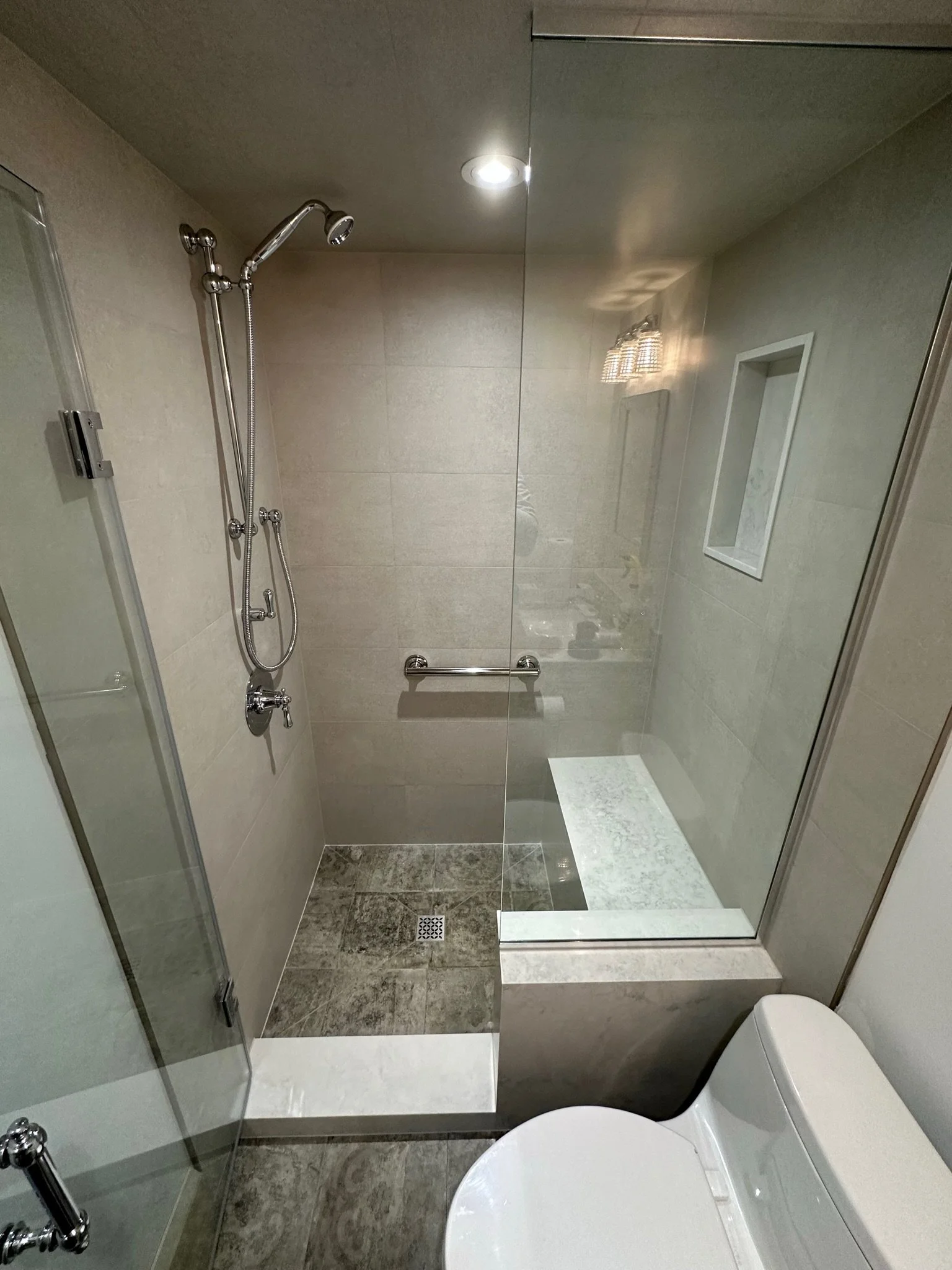 Bathroom with a walk-in shower, beige tiled walls, a chrome showerhead, a small recessed shelf, a white toilet, and a mirror reflecting the ceiling light.