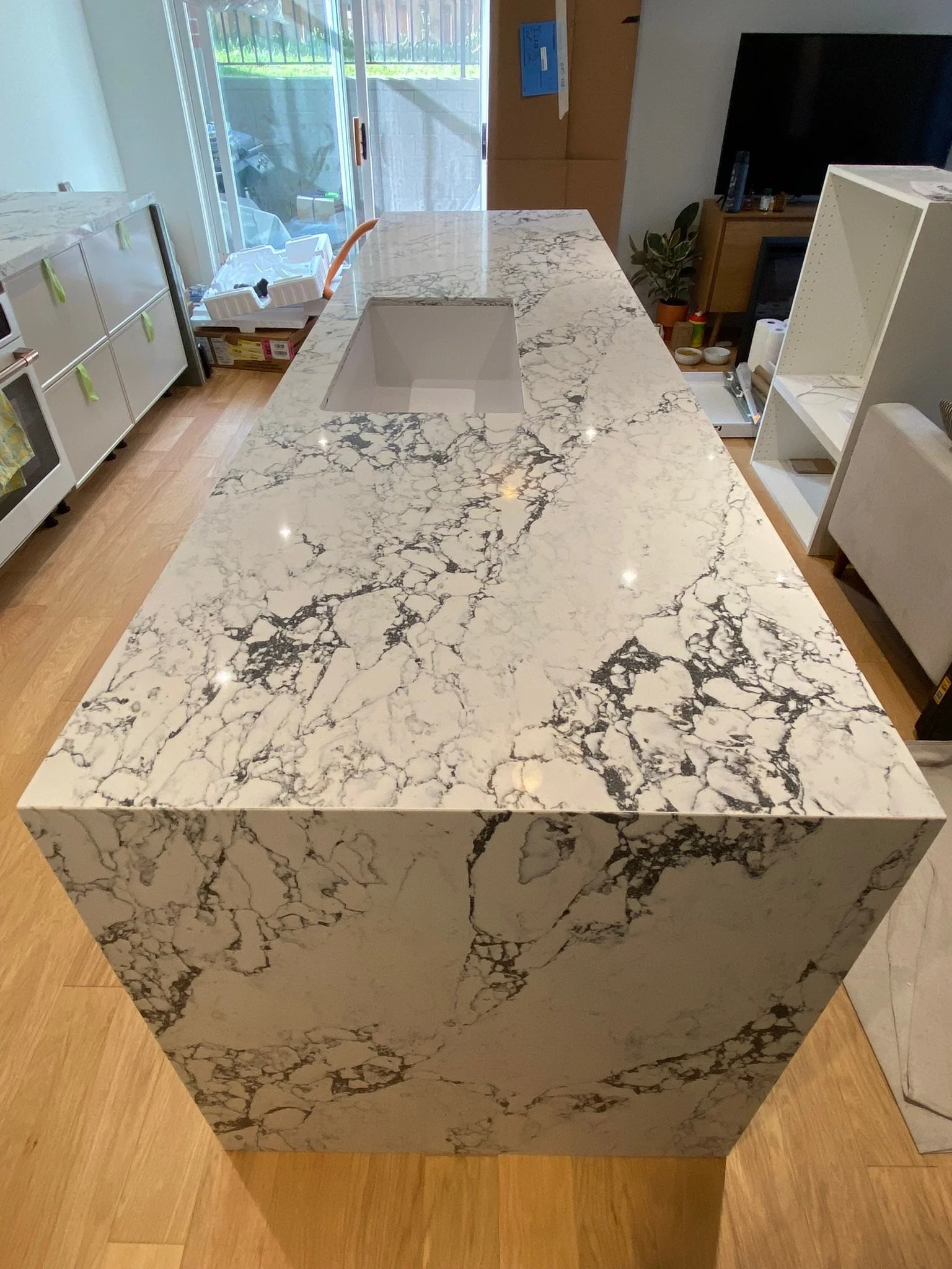 A large rectangular kitchen island with a white marble countertop featuring black veining. The island has a built-in sink and is situated in a kitchen with wooden flooring. There is a sliding glass door in the background, leading to an outdoor area with grass.