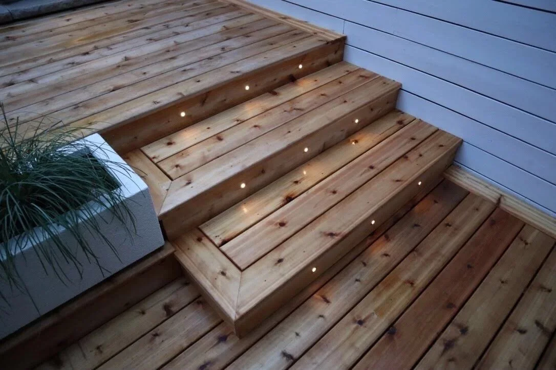 Wooden outdoor stairs with small built-in lights, next to a white planter with green plants, and a wall with horizontal siding.
