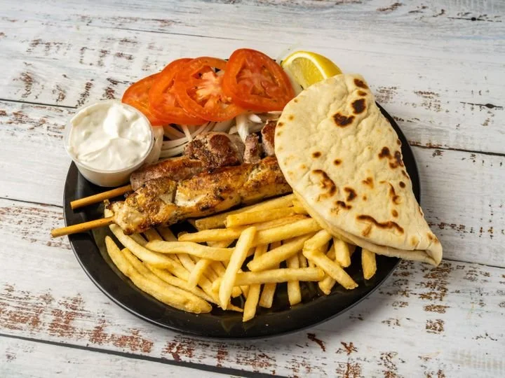 Plate of grilled chicken kebabs, French fries, pita bread, sliced tomatoes, onions, a lemon wedge, and a small bowl of yogurt sauce on a rustic wooden table.