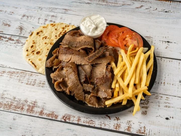Plate of grilled meat, French fries, sliced tomatoes, flatbread, and a small bowl of yogurt or sour cream on a rustic white wooden table.
