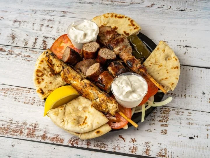 Mediterranean platter with grilled meat skewers, pita bread, hummus, tzatziki, and lemon wedges on a black plate