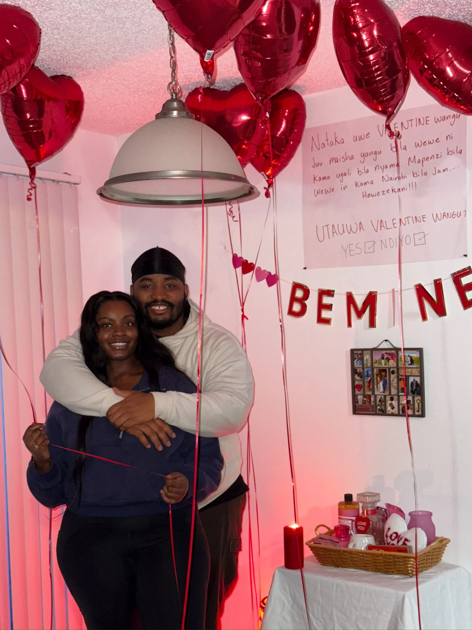 Two people hugging, surrounded by red heart-shaped balloons, with a Valentine's Day themed display in the background including a basket of gifts, candles, and Valentine's Day decorations.