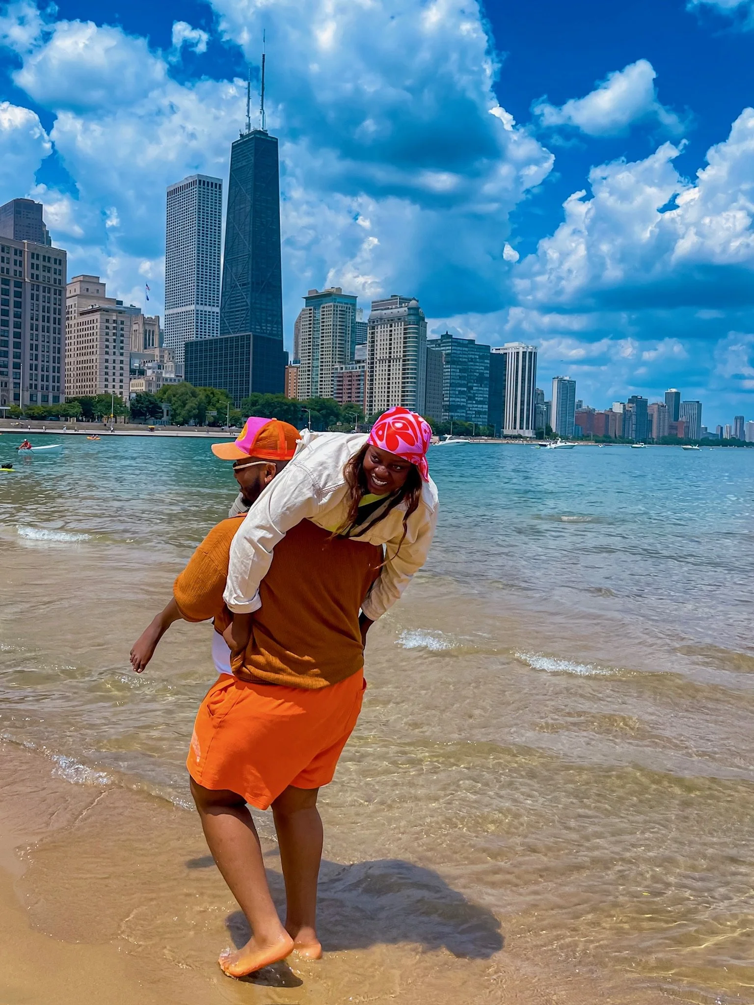 A woman with a red and white bandana, smiling, carrying a child on her back, standing in shallow water at a beach with a city skyline and tall buildings in the background under a partly cloudy sky.