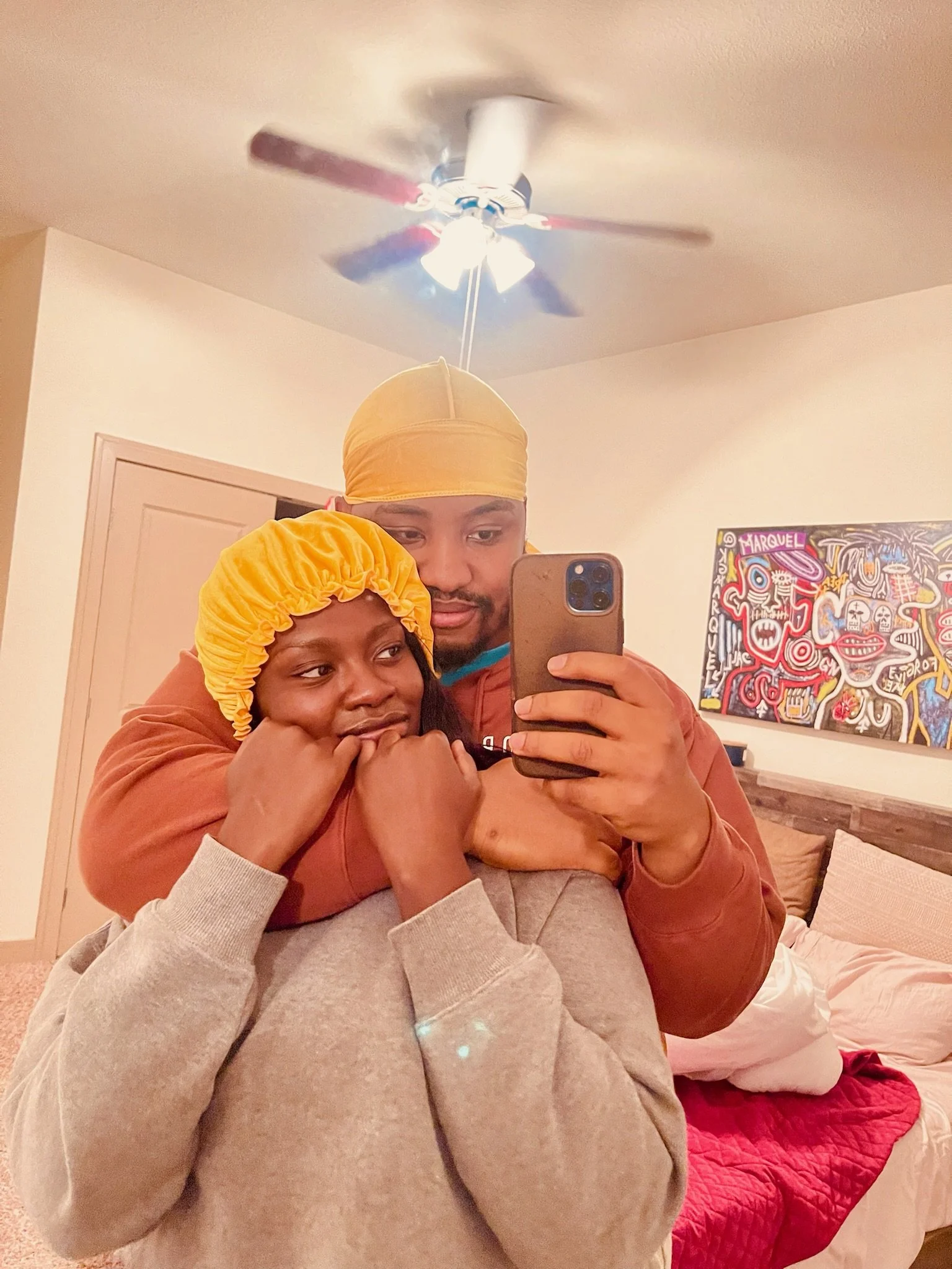 A man and a woman taking a mirror selfie inside a bedroom, with the woman wearing a yellow satin bonnet, and the man holding the phone to capture the photo.