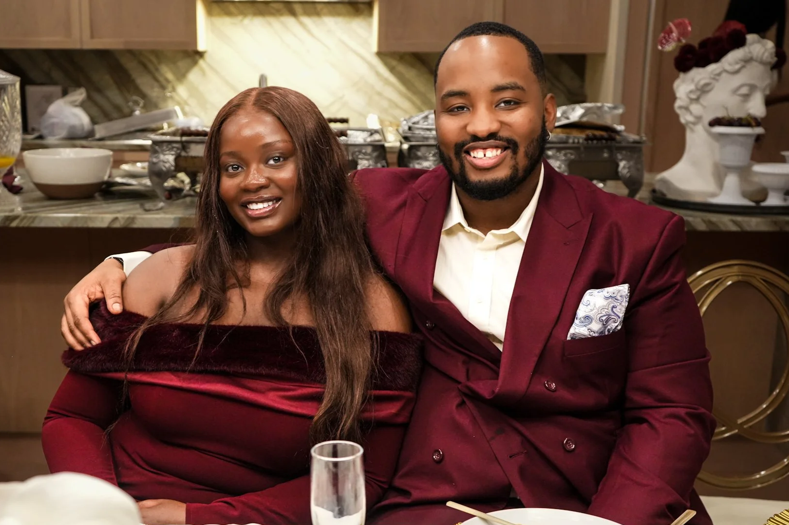 A smiling woman and man sitting closely together at a dinner event, with the woman wearing a maroon off-shoulder dress and the man in a maroon suit jacket and white shirt. The background features a buffet table and decorative sculptures.