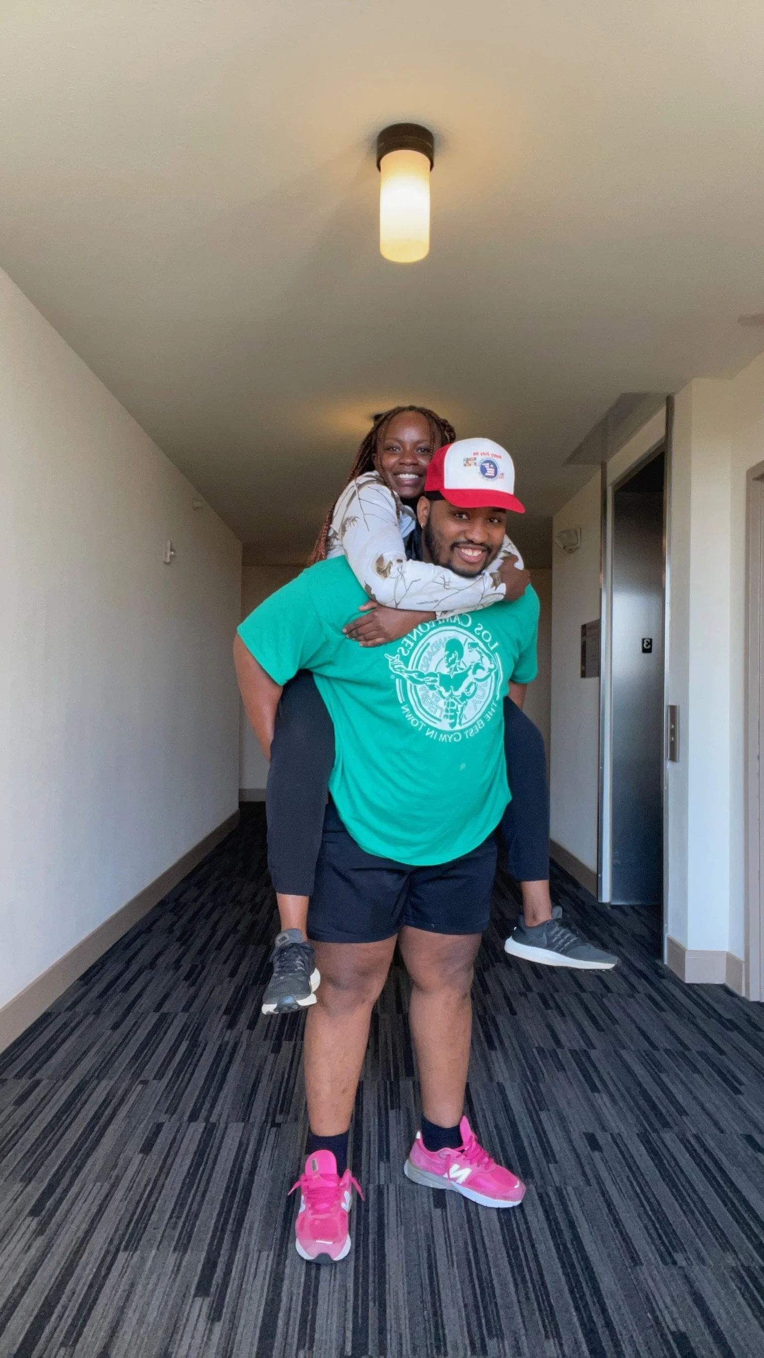 A man carrying a woman on his shoulders in an indoor hallway, both smiling and looking at the camera.