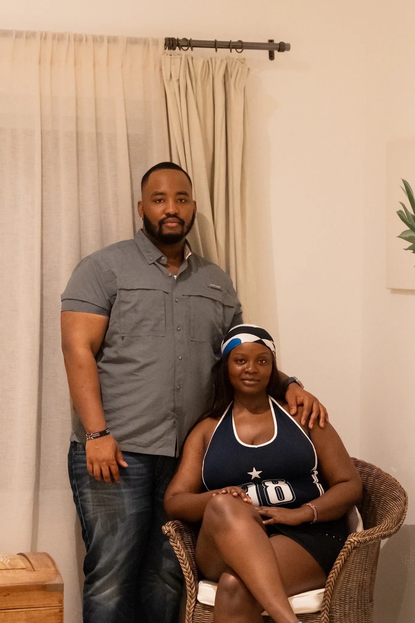 A man and a woman posing indoors, with the man standing and the woman sitting on a wicker chair, wearing a sports jersey and a headscarf.