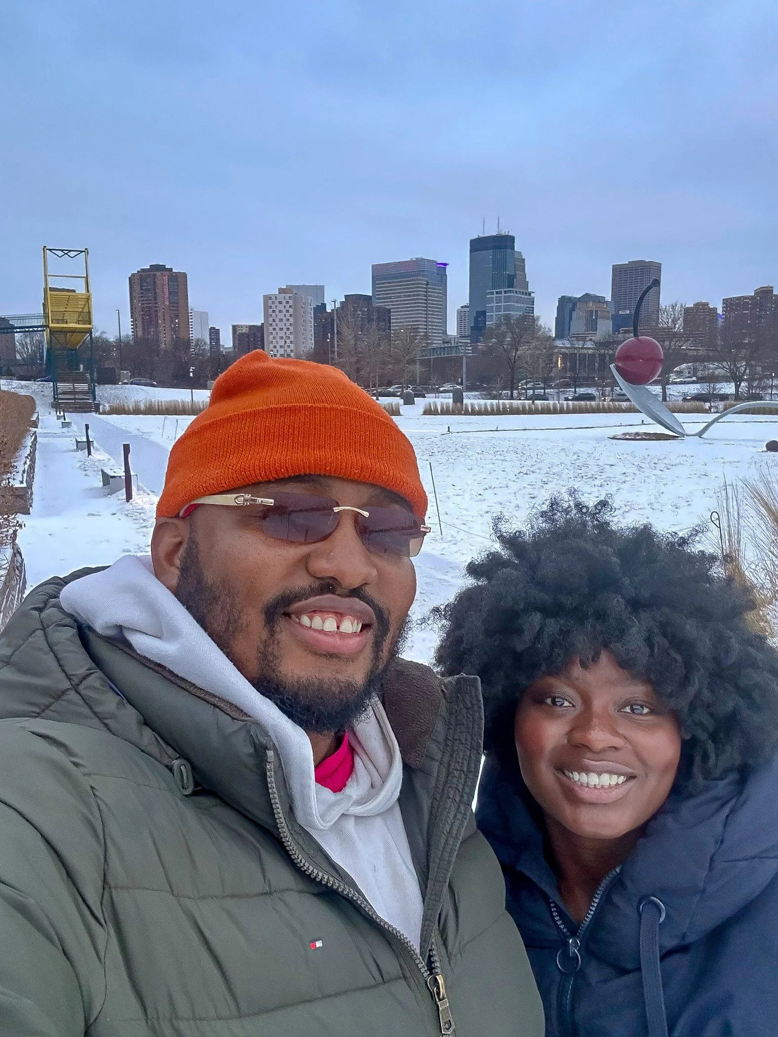 Two smiling people taking a selfie outdoors in a snowy urban park with a city skyline in the background, wearing winter jackets and hats.
