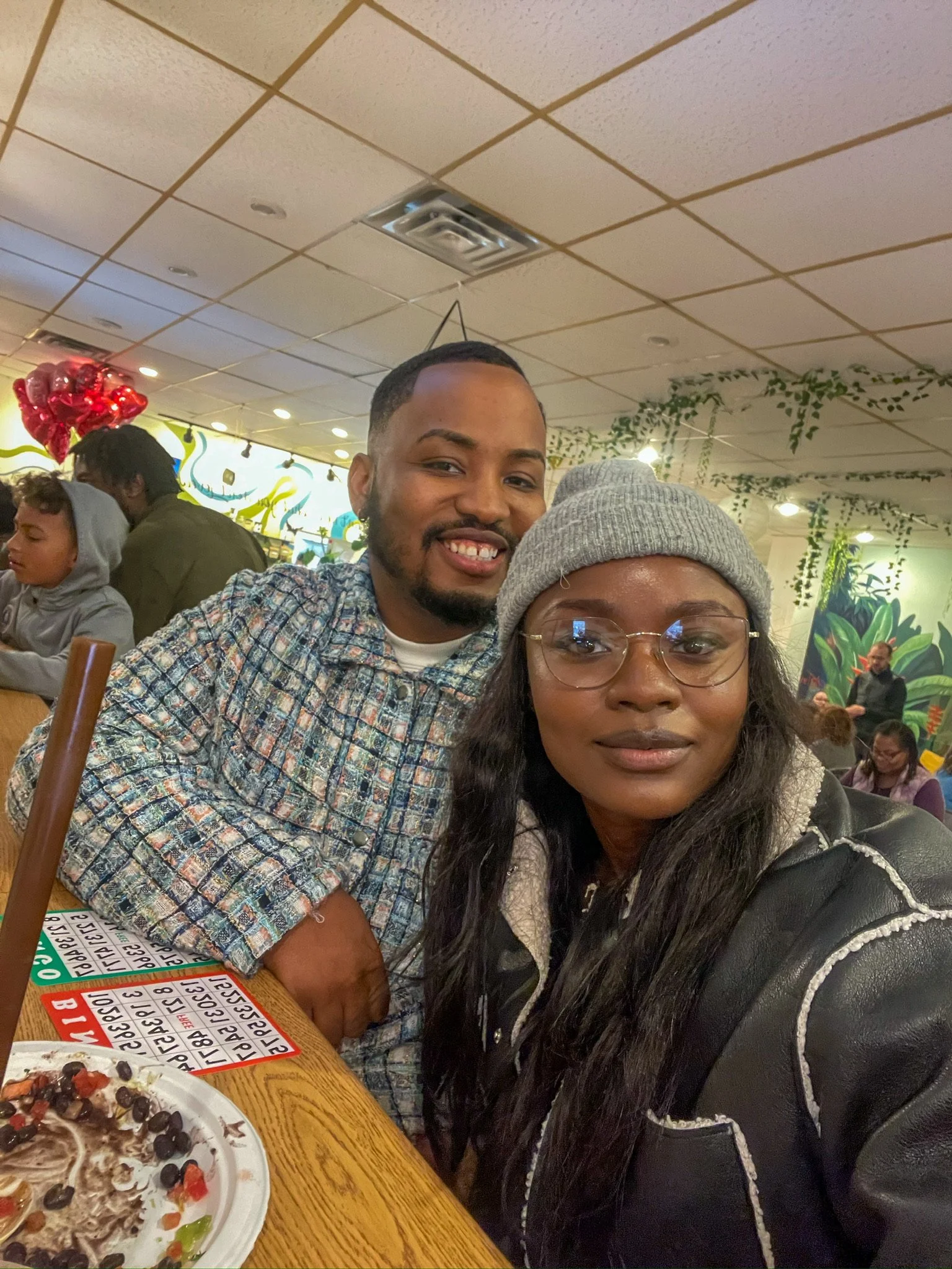 Two friends taking a selfie inside a restaurant with a vibrant, tropical-themed decor, with a plate of food and a bingo sheet visible on the table.