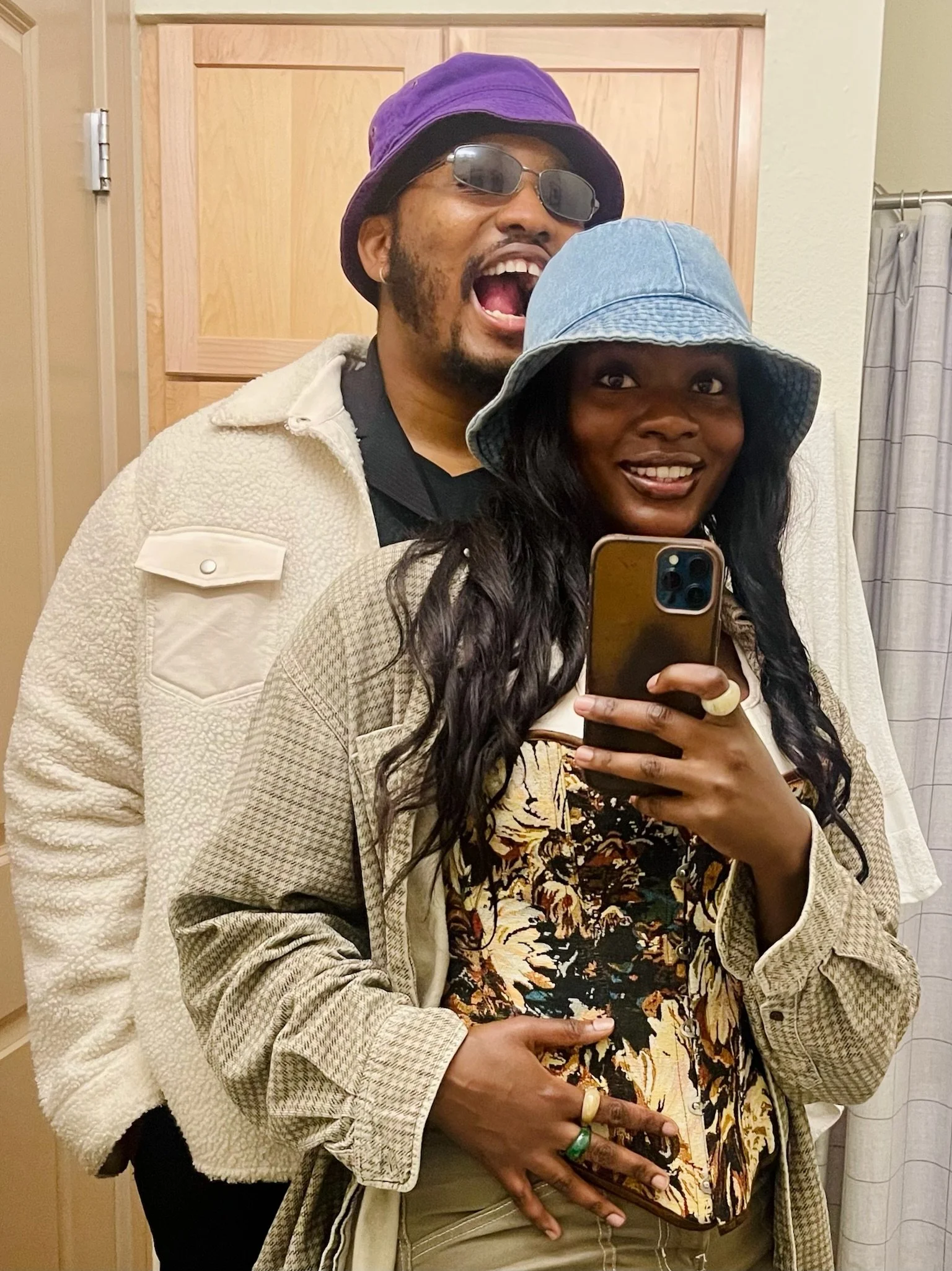 A couple taking a mirror selfie in a bathroom. The man is wearing sunglasses, a purple bucket hat, a beige jacket, and has a beard. The woman is wearing a light blue hat, a patterned top, and a beige jacket, holding a smartphone with a gold case. The