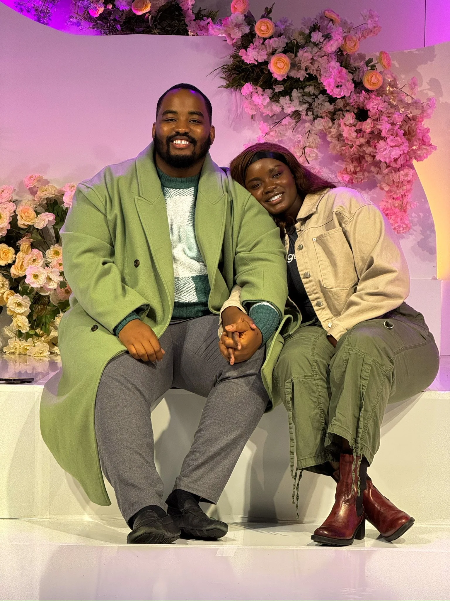 Two people sitting and holding hands, with pink floral arrangements behind them, in a setting with pink and purple lighting.