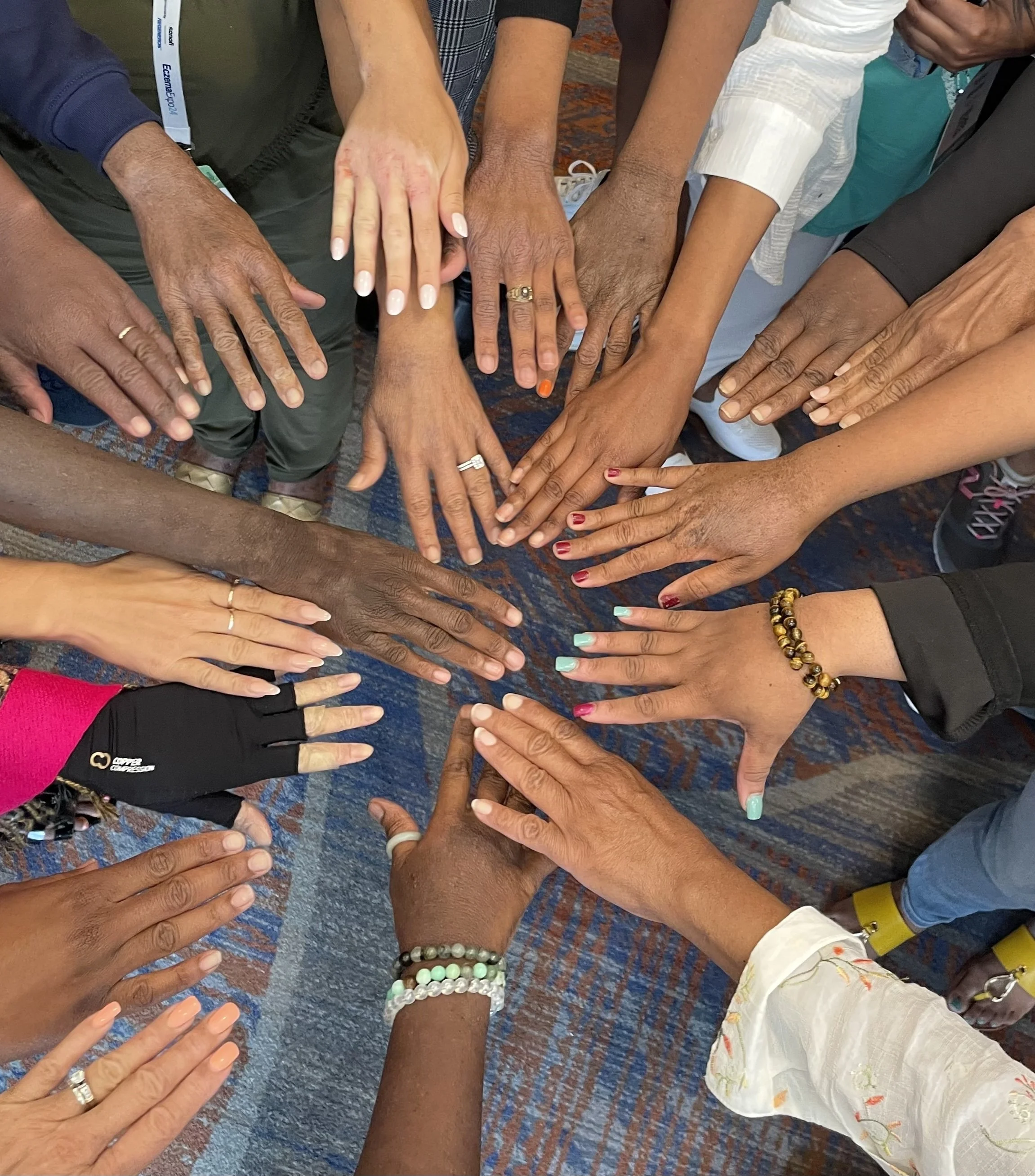 A group of diverse hands with different skin tones are stacked together in a circle, symbolizing unity and teamwork. Several hands are adorned with rings, bracelets, and nail polish, and they are positioned over a textured surface.