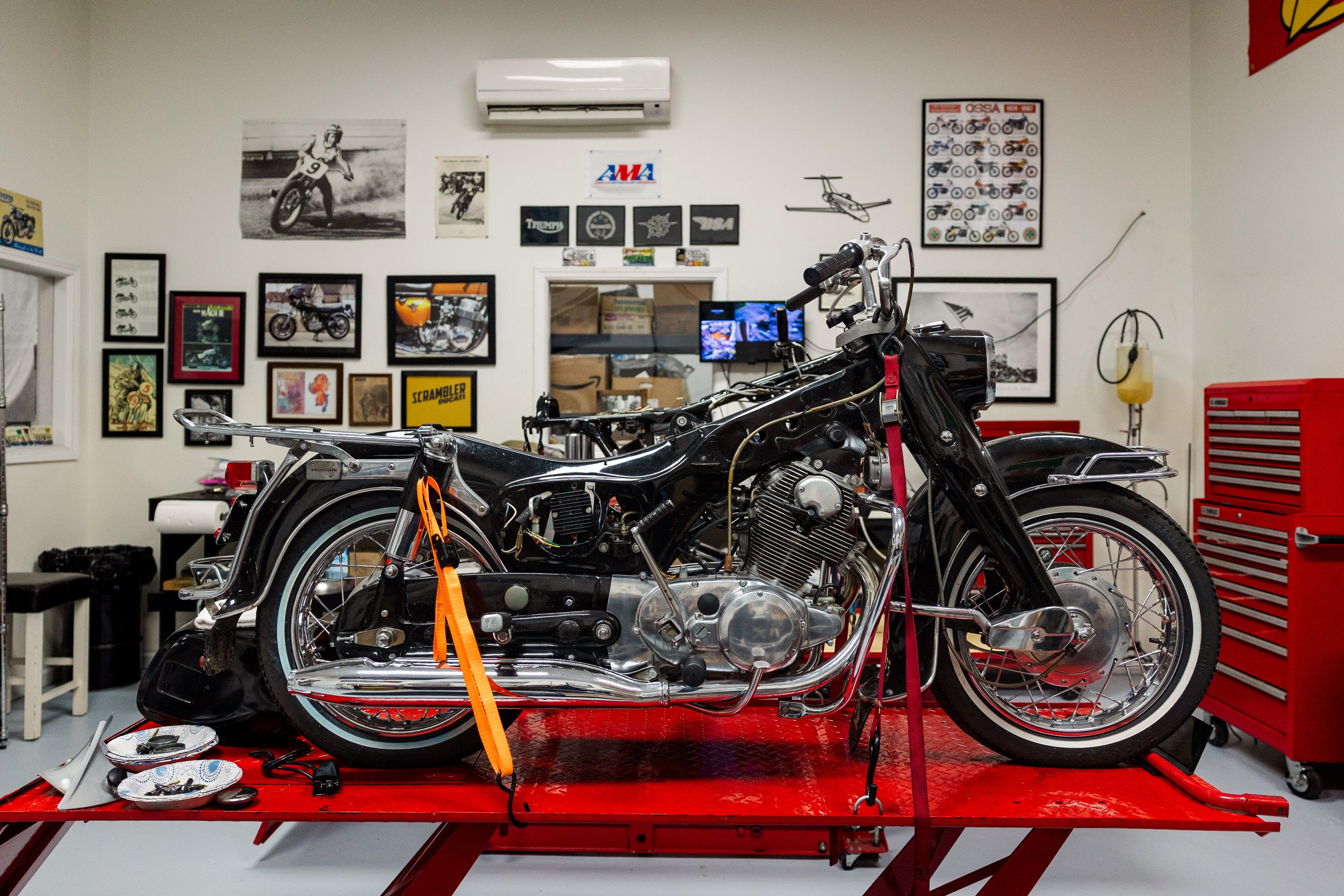 A motorcycle is on a red work table in a workshop, with tools and parts nearby. The workshop wall has various framed posters and pictures, including motorcycle-related images and logos, with a wall-mounted air conditioning unit at the top.