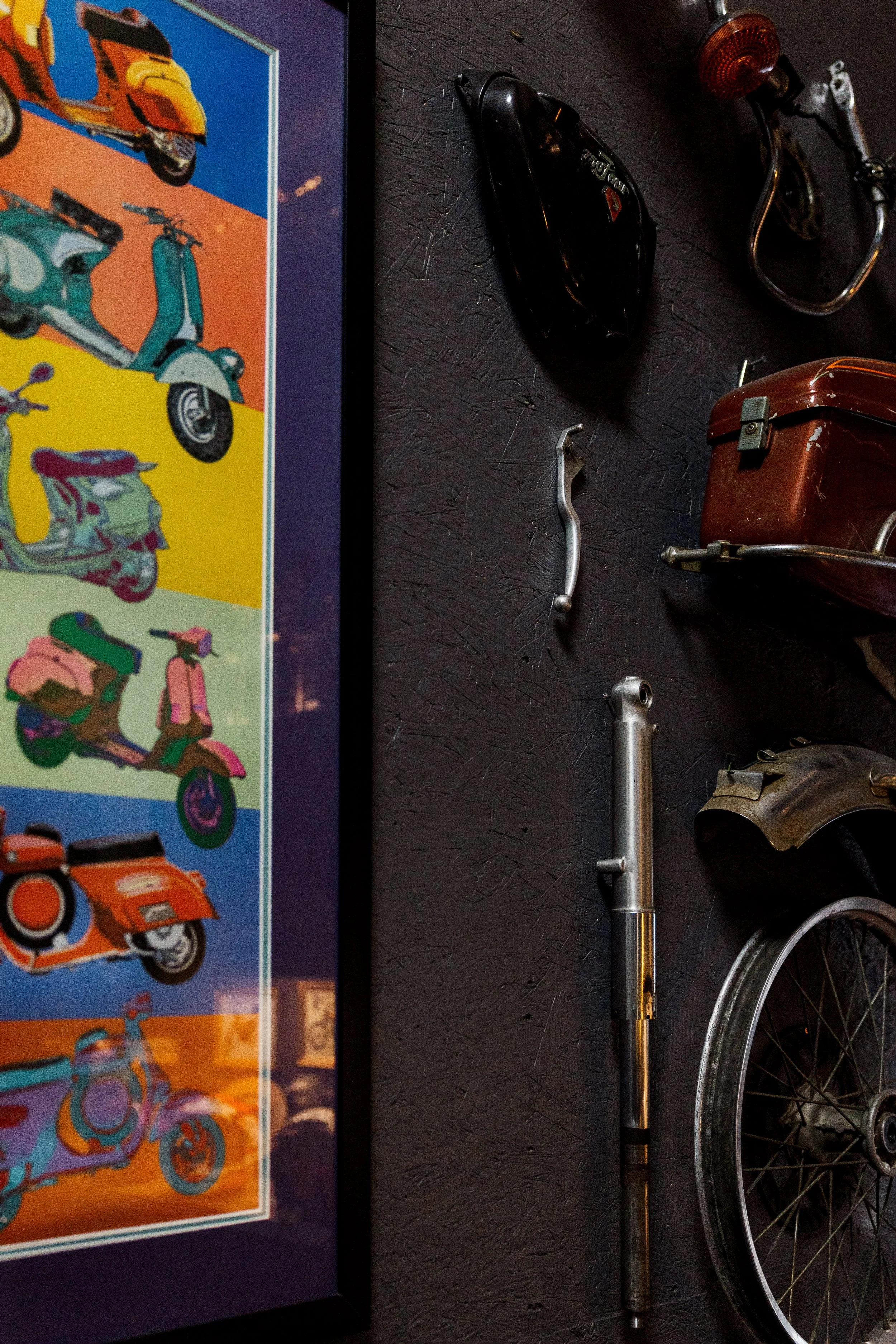 A black motorcycle handlebar with a brake lever, next to a rusted metallic part, laying on a black textured surface. There is also a black object, possibly a helmet, and a small orange reflector on a black cord. Inside Birmingham Vintage Cycle