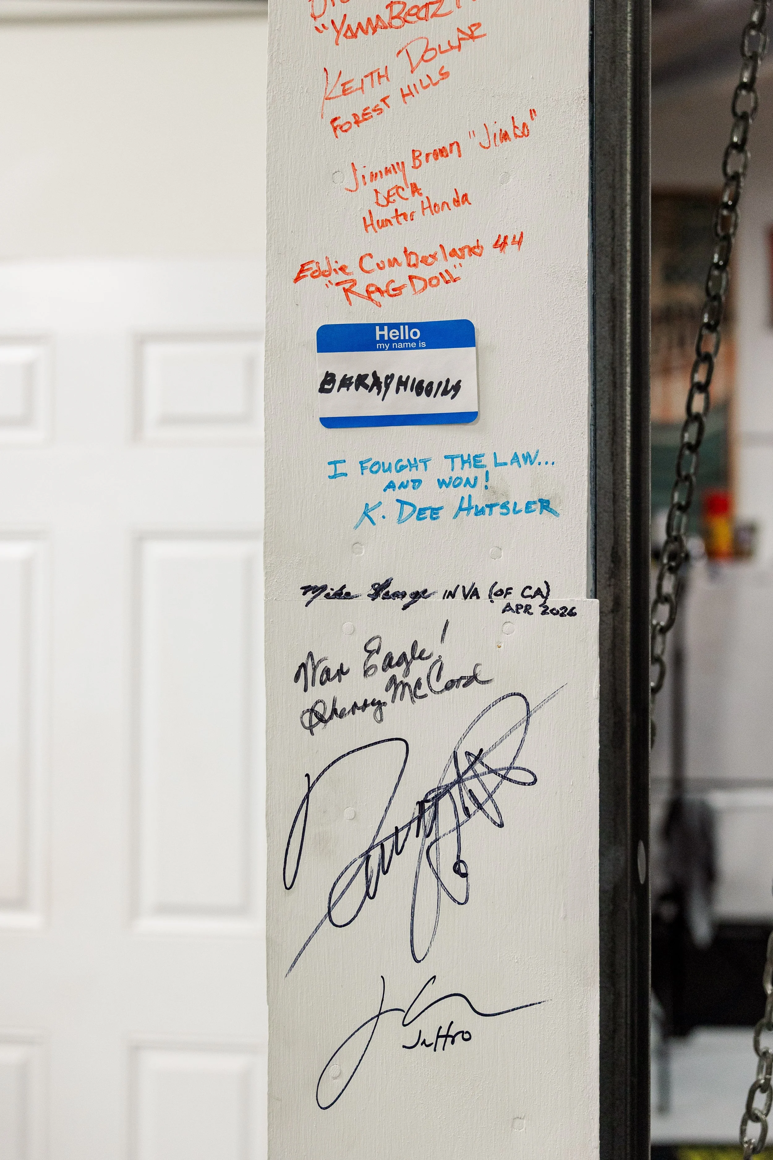 A wall with handwritten notes and signatures. There is a blue and white 'Hello my name is' badge with the name 'BARRY HIGGINS' in black marker. The notes mention various names and titles, including 'Jimmy Broom,' 'Eddie Cumbelands,' and a quote 'I fo