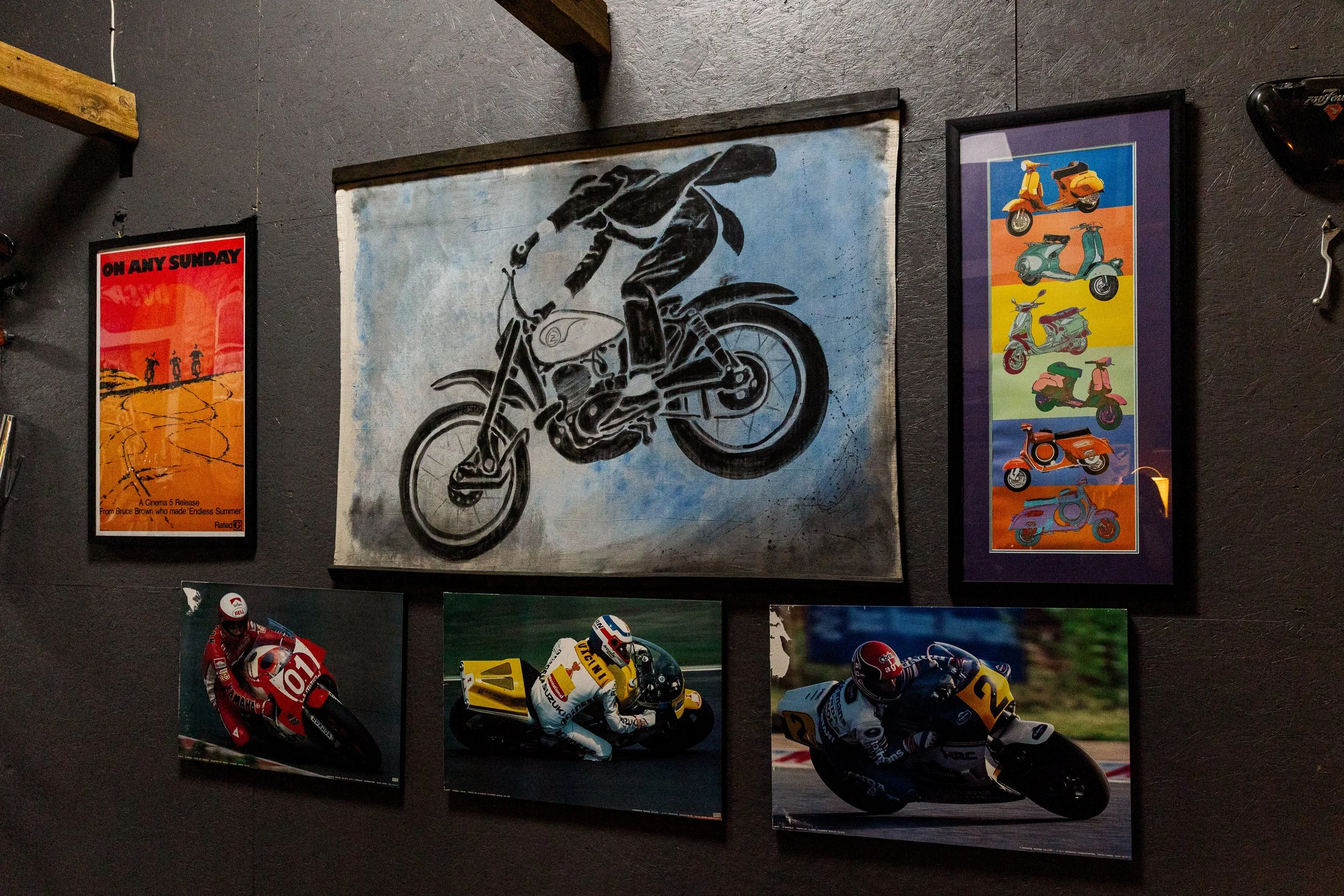 Various posters and artwork on a gray wall, including a black and white illustration of a person riding a motorcycle, and other colorful images of scooters, motorcycles, and racing scenes. Vintage motorcycle collection on display at BVC