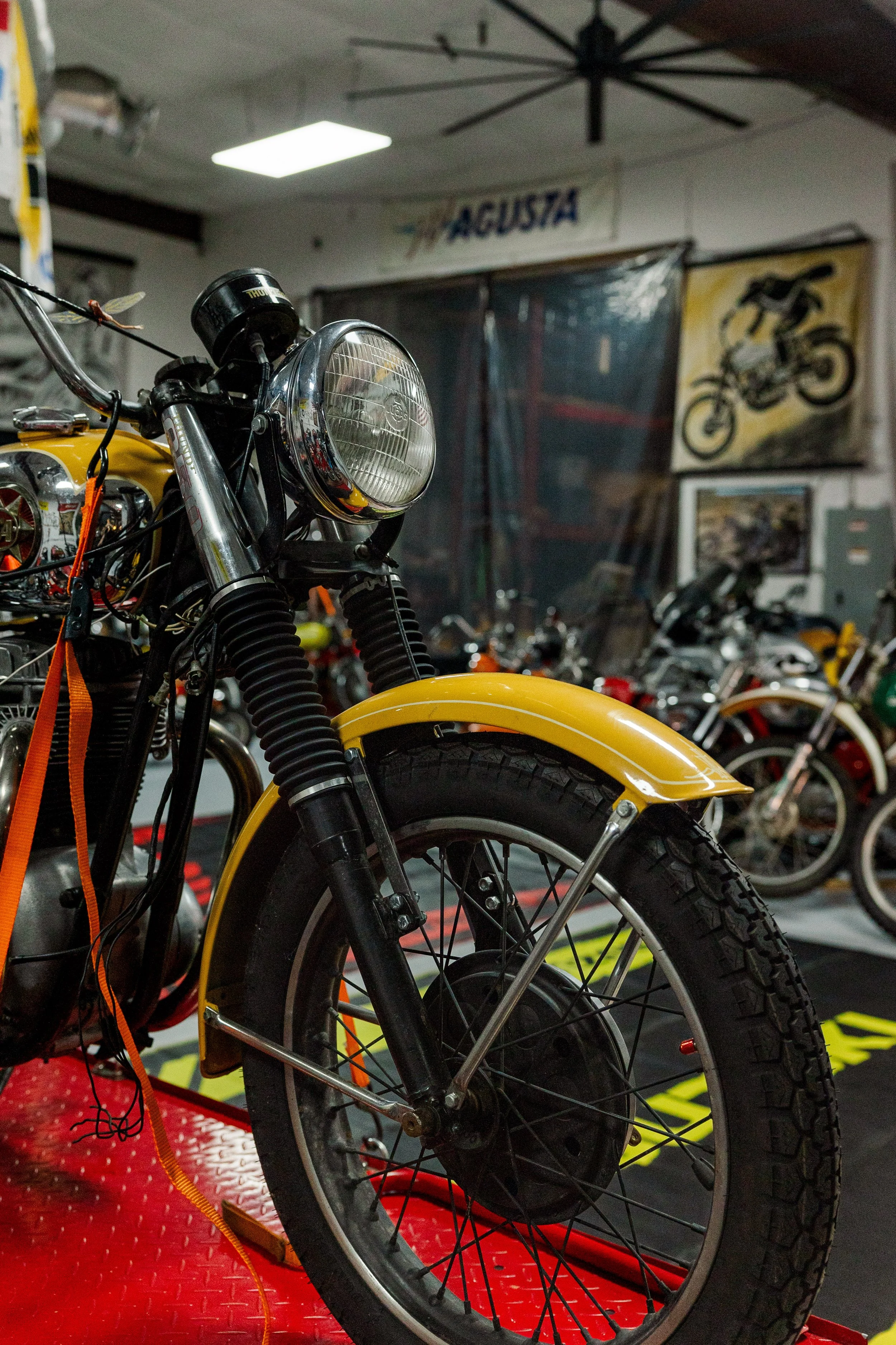Close-up of a vintage yellow motorcycle in a garage or workshop environment with various motorcycle posters and tools in the background. Vintage motorcycle collection on display at Birmingham Vintage Cycle in Birmingham Alabama