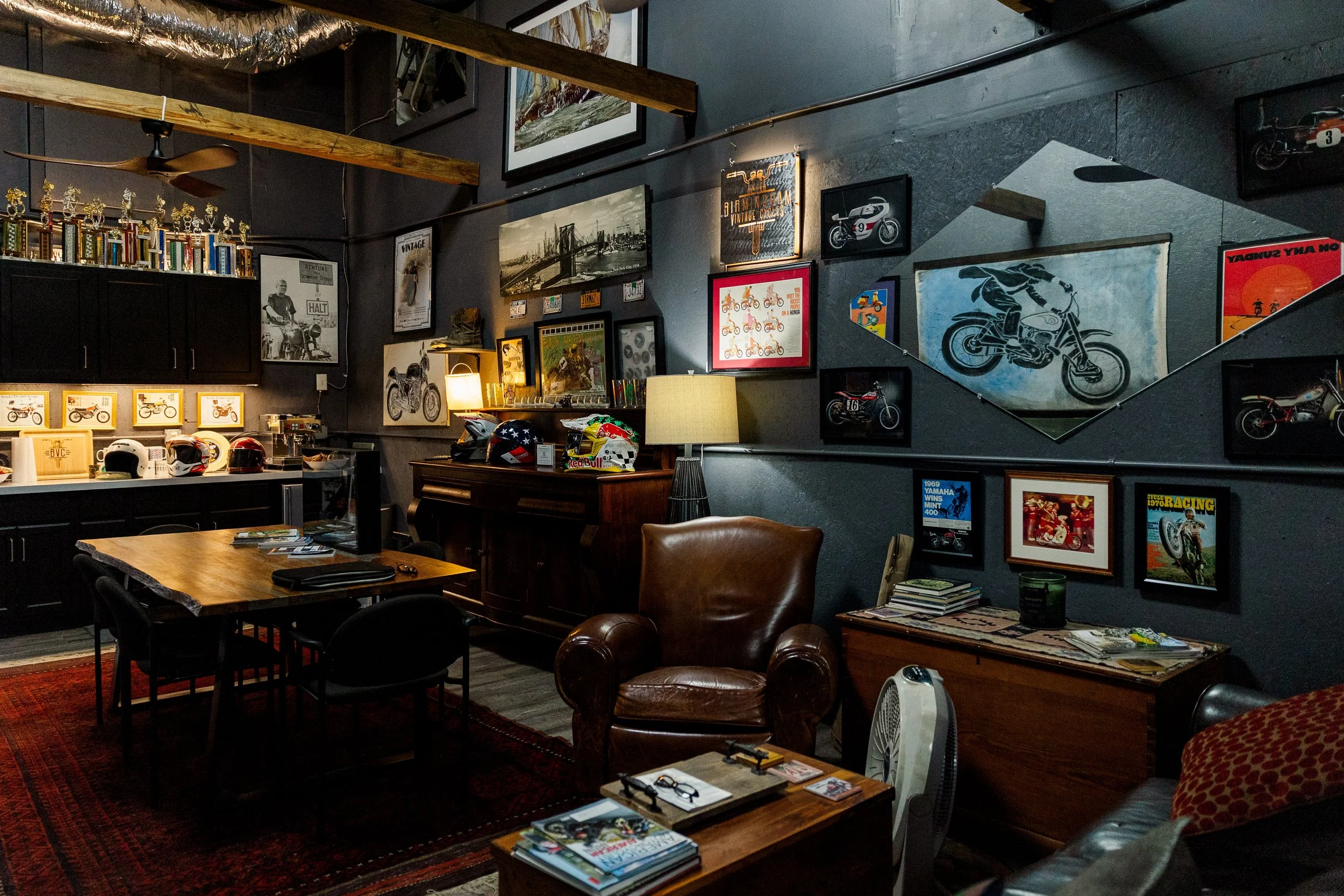 Inside Birmingham Vintage Cycle, a vintage motorcycle shop. There are ledges and wall art featuring motorcycles, racing posters, and memorabilia, with a table and chairs in the foreground and various motorcycle helmets on the table and shelf.