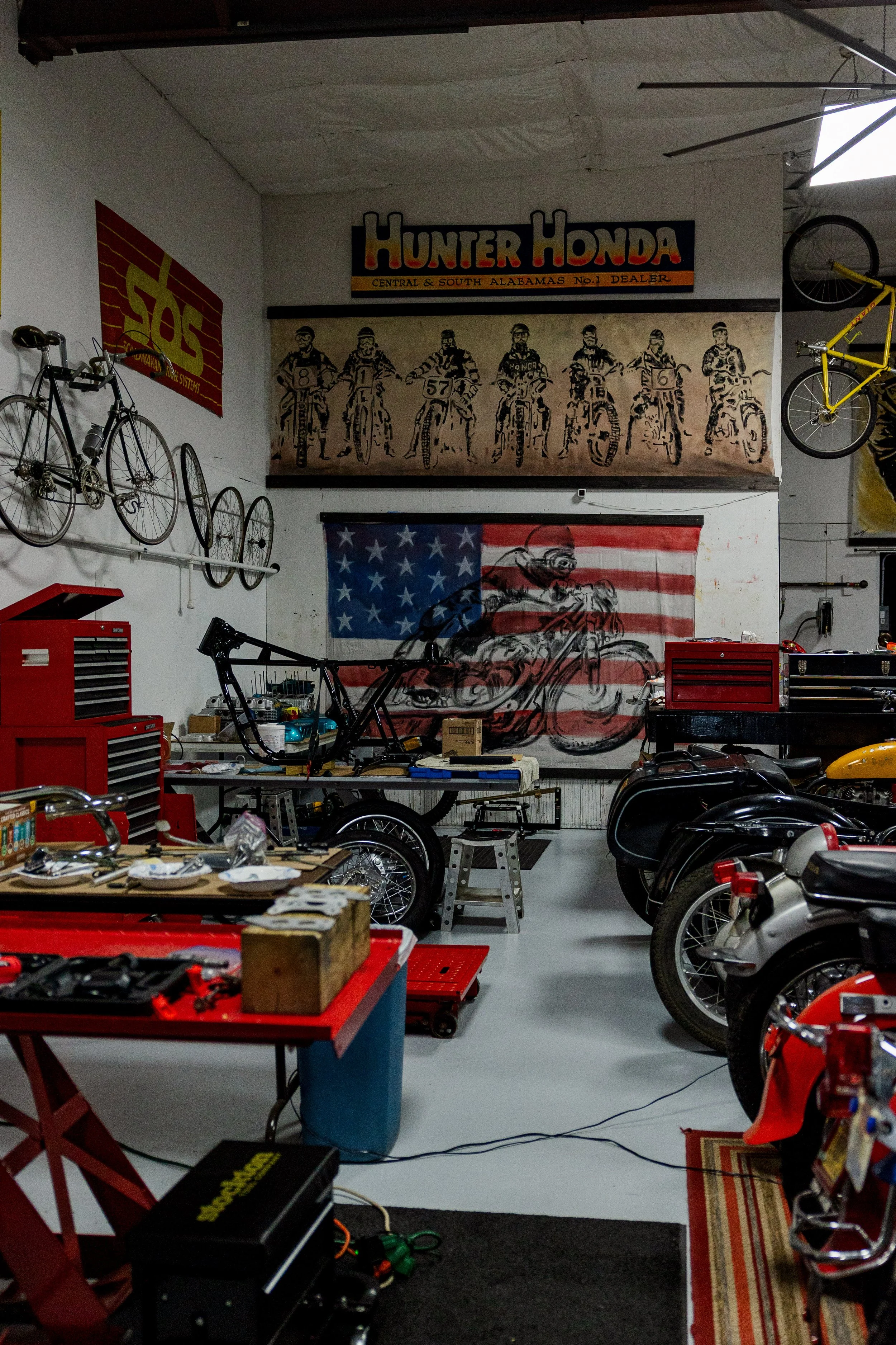 The image shows a motorcycle repair or customization workshop with motorcycles, tools, and vintage motorcycle artwork. There are bicycles mounted on the wall, a red workbench with tools, and a large American flag mural with a rider and motorcycle por