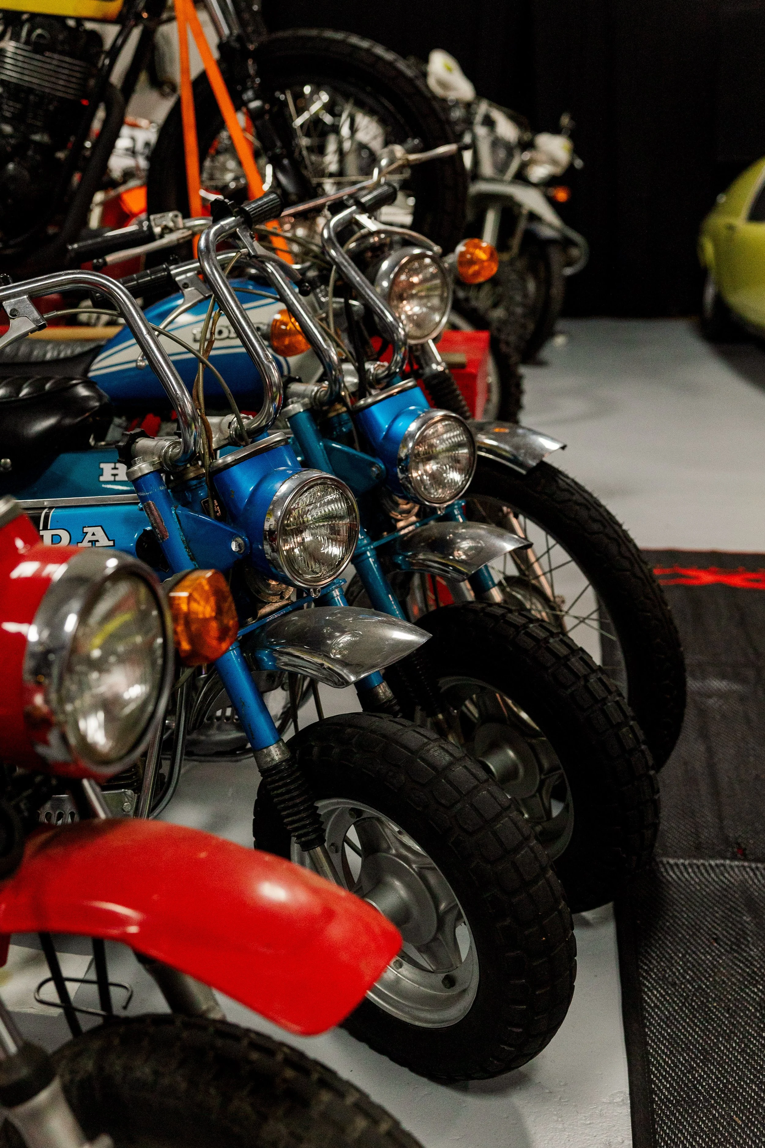 Multiple vintage motorcycles with large tires lined up indoors, featuring bright blue and red colors, with some motorcycles showing chrome and headlight details. Vintage motorcycle collection on display at Birmingham Vintage Cycle in Birmingham