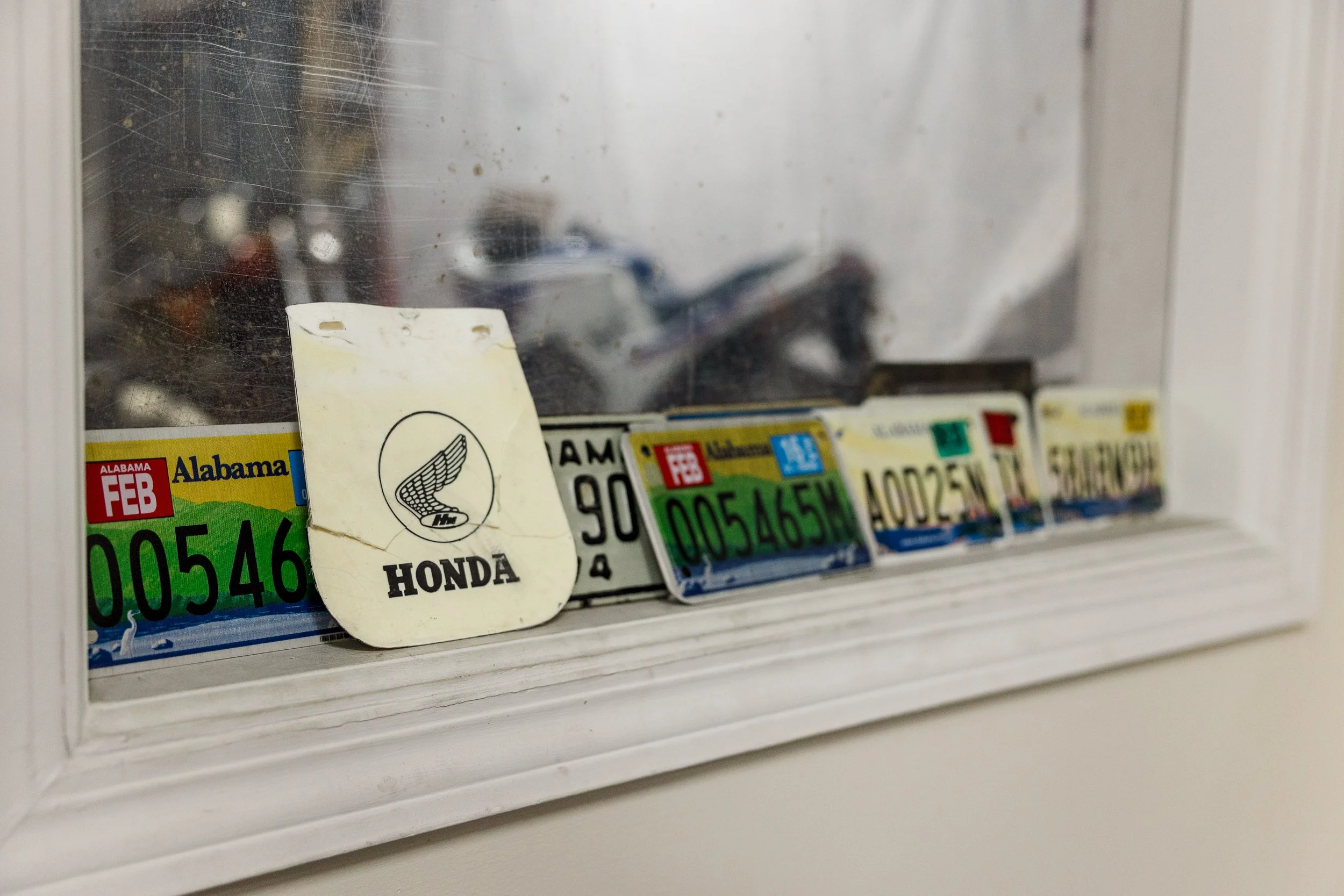 Close-up of a collection of used vehicle license plates behind a dirty glass window, with a small, yellowed paper tag with black writing and a logo attached to the display. Inside Birmingham Vintage Cycle, a vintage motorcycle shop and community