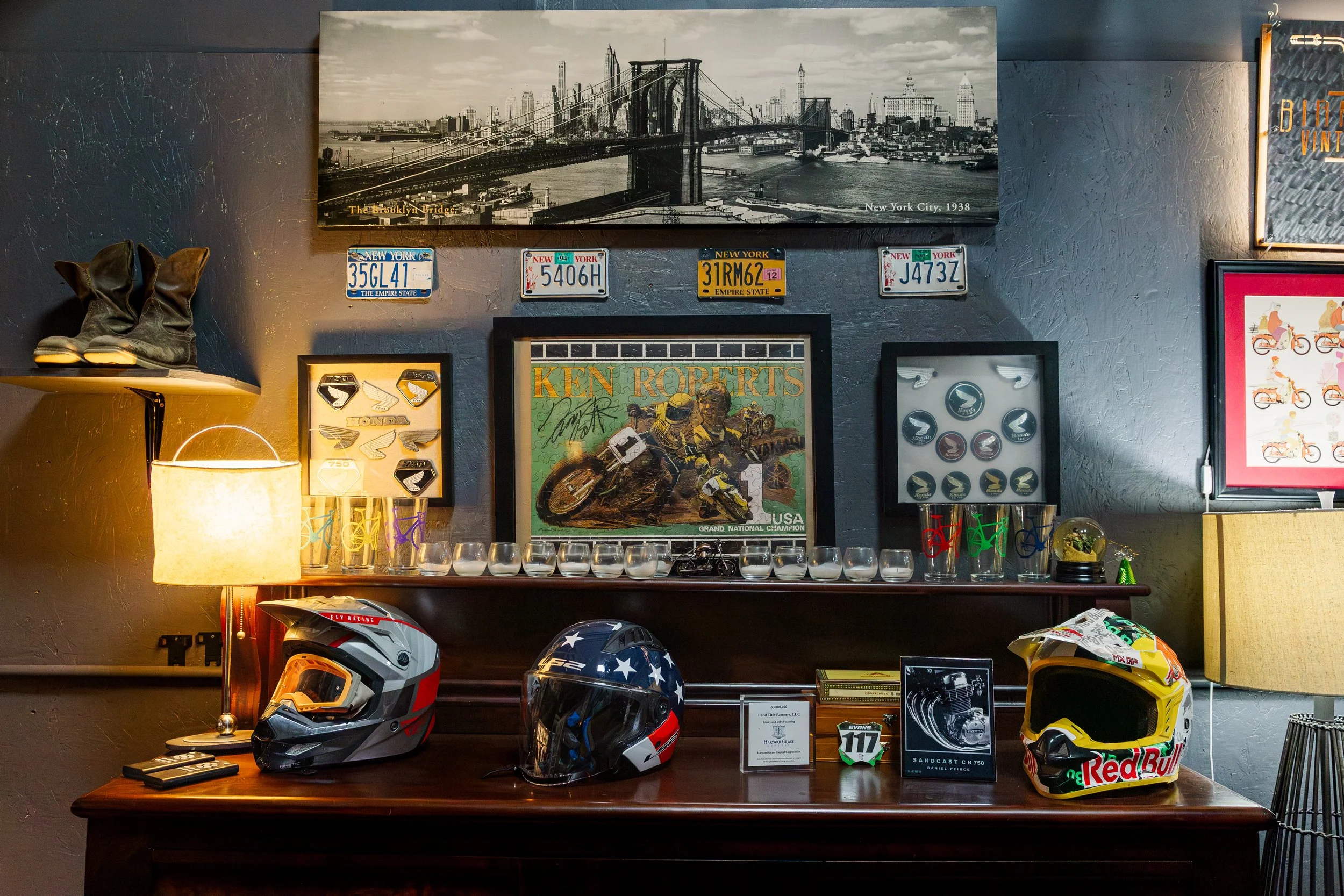 Display of motorcycle Helmets on a wooden table, framed racing-related posters, vintage license plates on a dark textured wall, and a black and white photo of Brooklyn Bridge and New York City skyline from 1938. Inside Birmingham Vintage Cycle