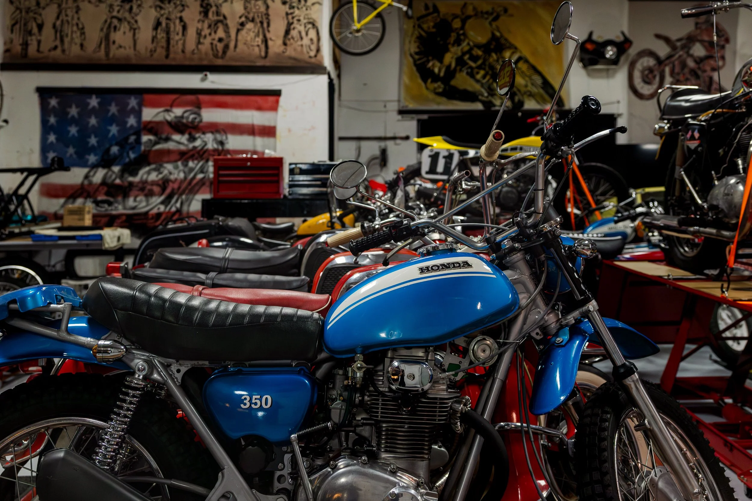 A vintage blue Honda motorcycle with a black leather seat and chrome details in a garage filled with various bikes, tools, and artwork. Vintage motorcycle collection on display at Birmingham Vintage Cycle in Birmingham Alabama
