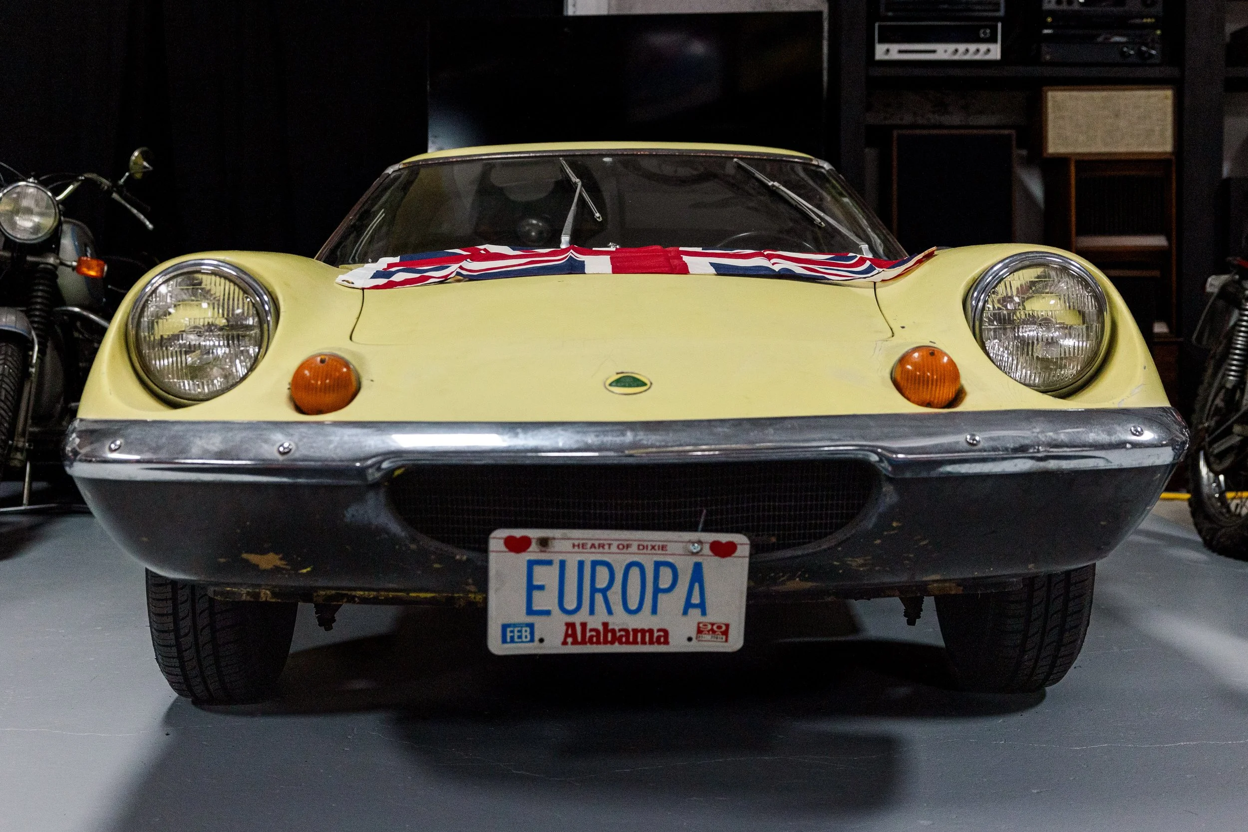 Yellow vintage Lotus Elise sports car with a Union Jack flag on the front hood, parked indoors with other vehicles and machinery in the background. Vintage motorcycle collection on display at Birmingham Vintage Cycle in Birmingham Alabama