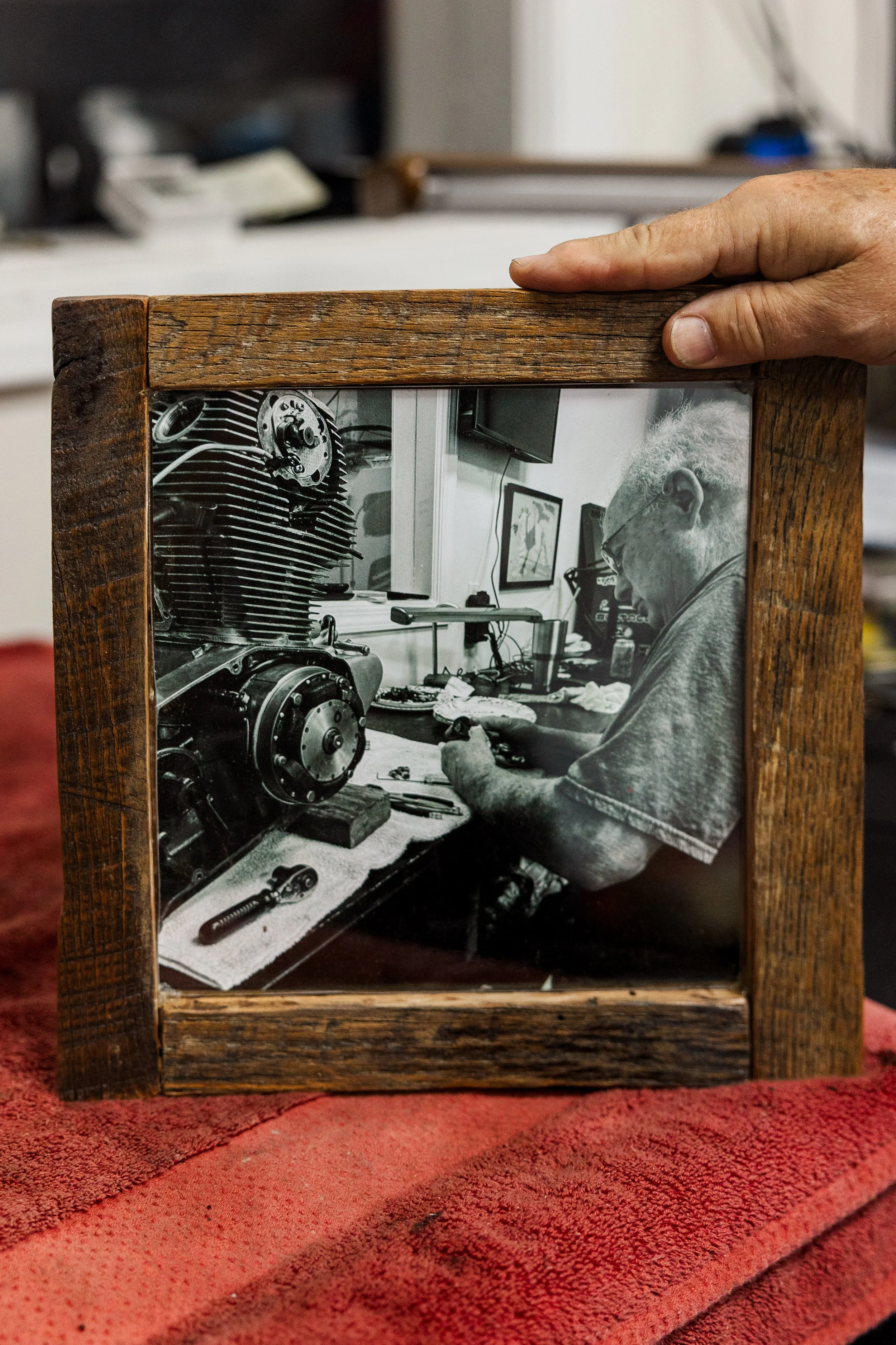 A person with gray hair and glasses working on a mechanical engine inside a room, as seen through a framed photo. Inside Birmingham Vintage Cycle, a vintage motorcycle shop and community space in Birmingham Alabama