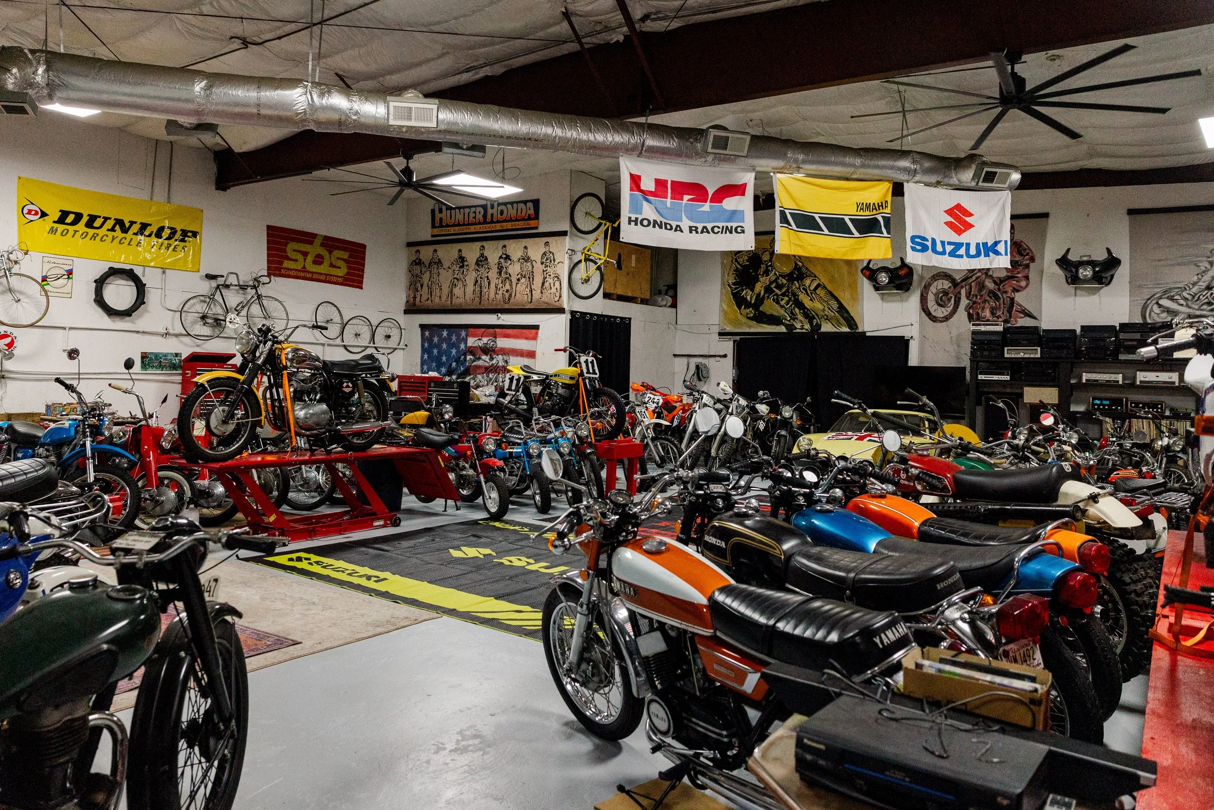 Interior of a vintage motorcycle and bicycle collection with various bikes and memorabilia, including flags for Dunlop, Hunter Honda, Honda Racing, Yamaha, and Suzuki, and a row of large stereo speakers. Vintage motorcycle collection on display BVC