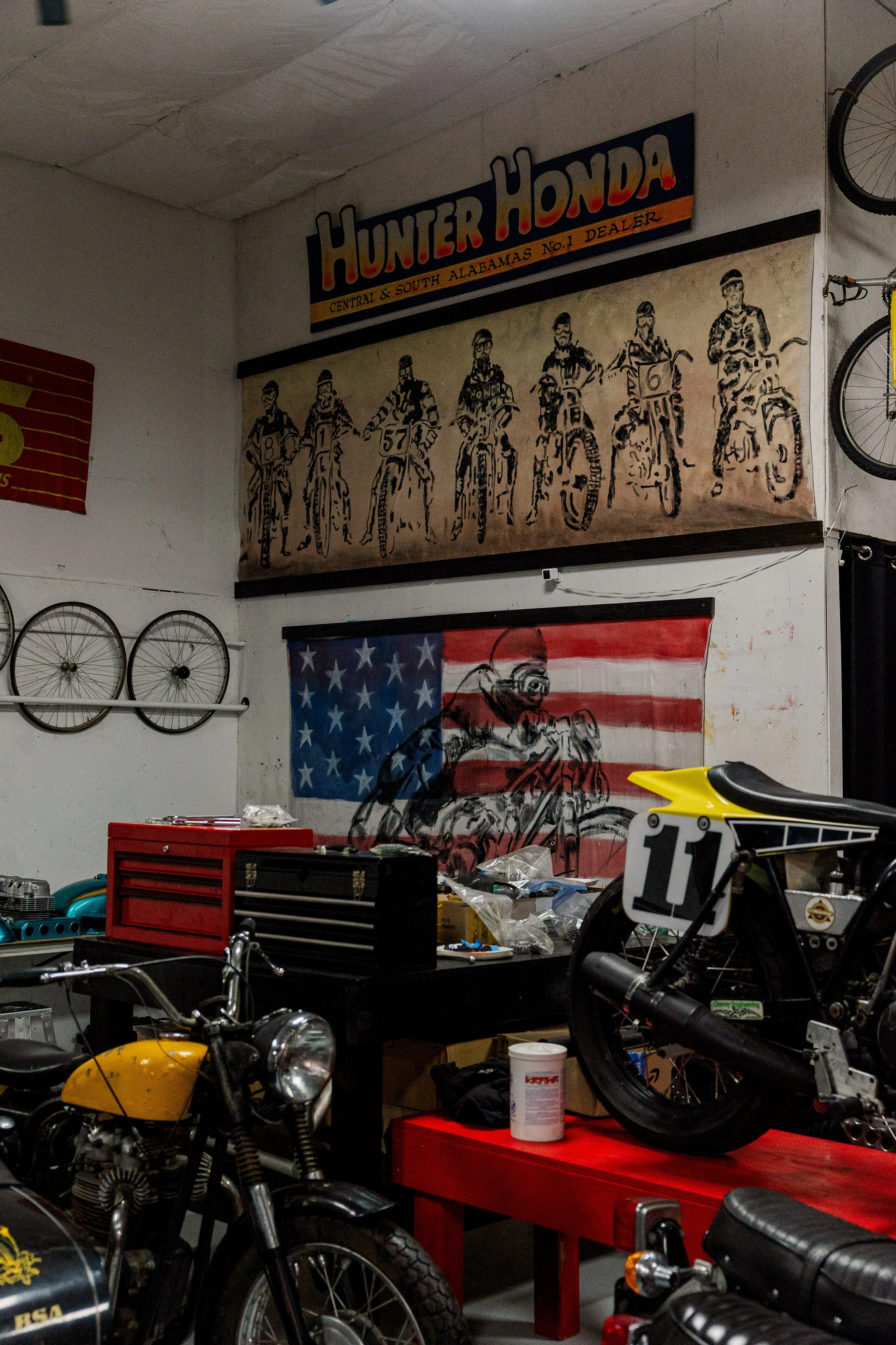 Indoor workshop filled with vintage motorcycles, tools, and parts. A banner on the wall reads "Hunter Honda," with a photo of motorcycle riders below it. Items are scattered on tables and shelves. Vintage motorcycle collection on display 