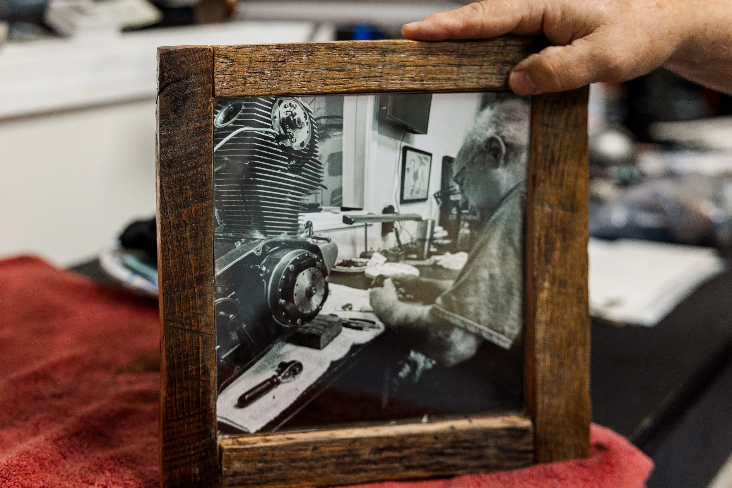 A person holding a wooden-framed black and white photograph of an engine workbench with tools and machinery. Inside Birmingham Vintage Cycle, a vintage motorcycle shop and community space in Birmingham Alabama