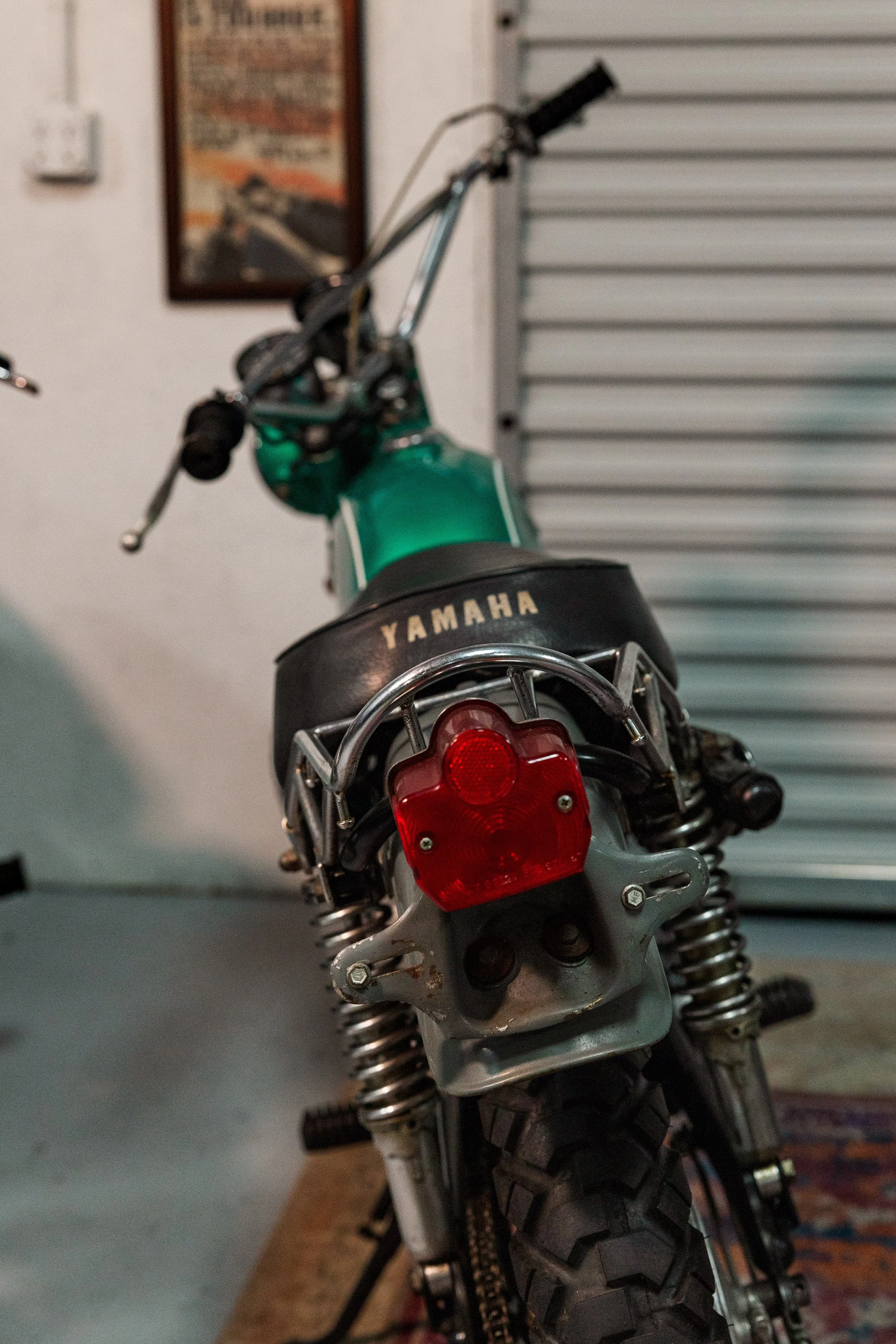 A close-up of a Yamaha dirt bike's rear, showing the tire, suspension, and red rear light, with the front unassembled handlebar and a roller door in the background. Vintage motorcycle collection on display at Birmingham Vintage Cycle in Birmingham
