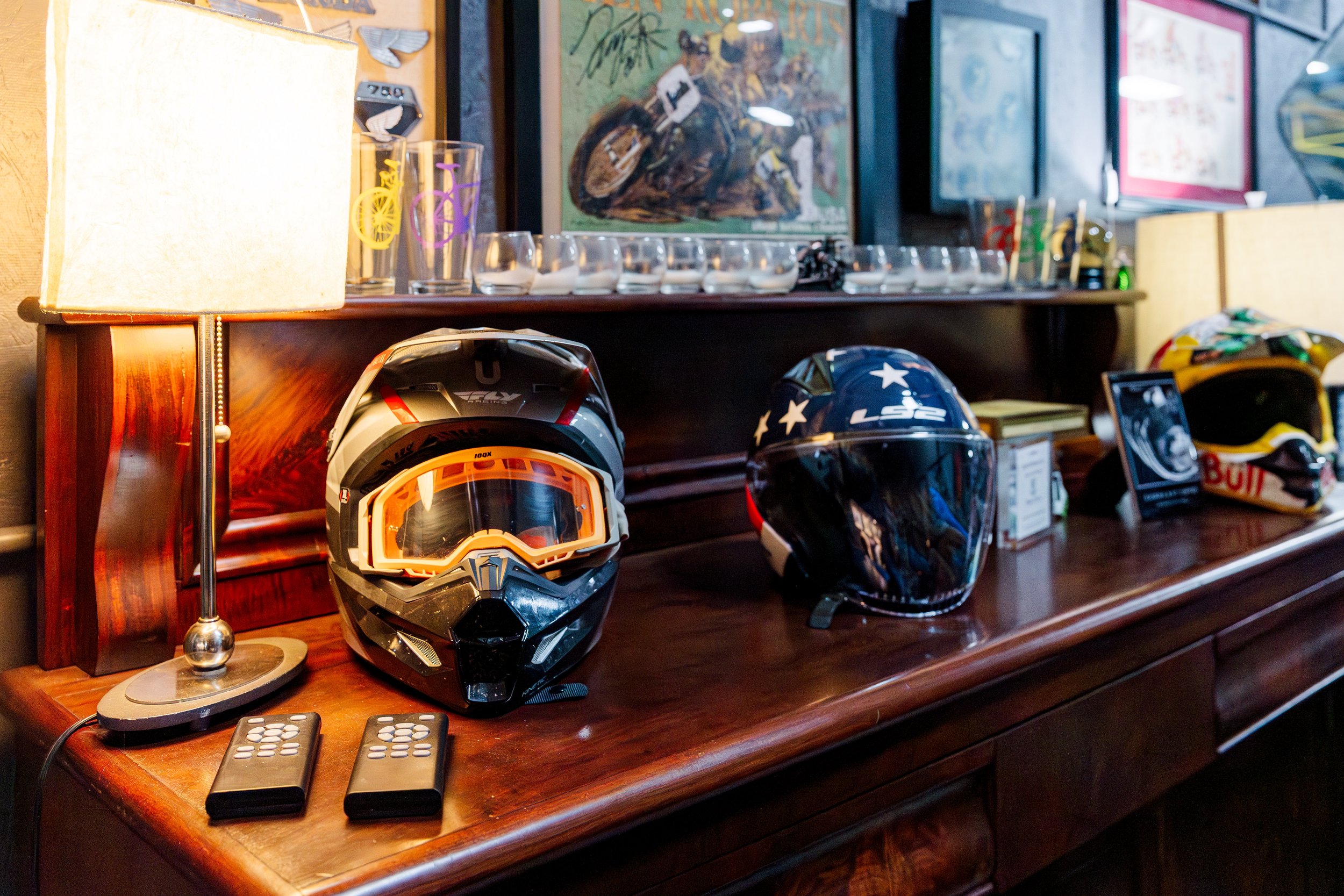 A wooden dresser with three motorcycle helmets on top. One helmet is black with yellow and red accents, the second is black with white stars, and the third is yellow and black. A table lamp with a beige shade and two TV remotes are also on the dresse