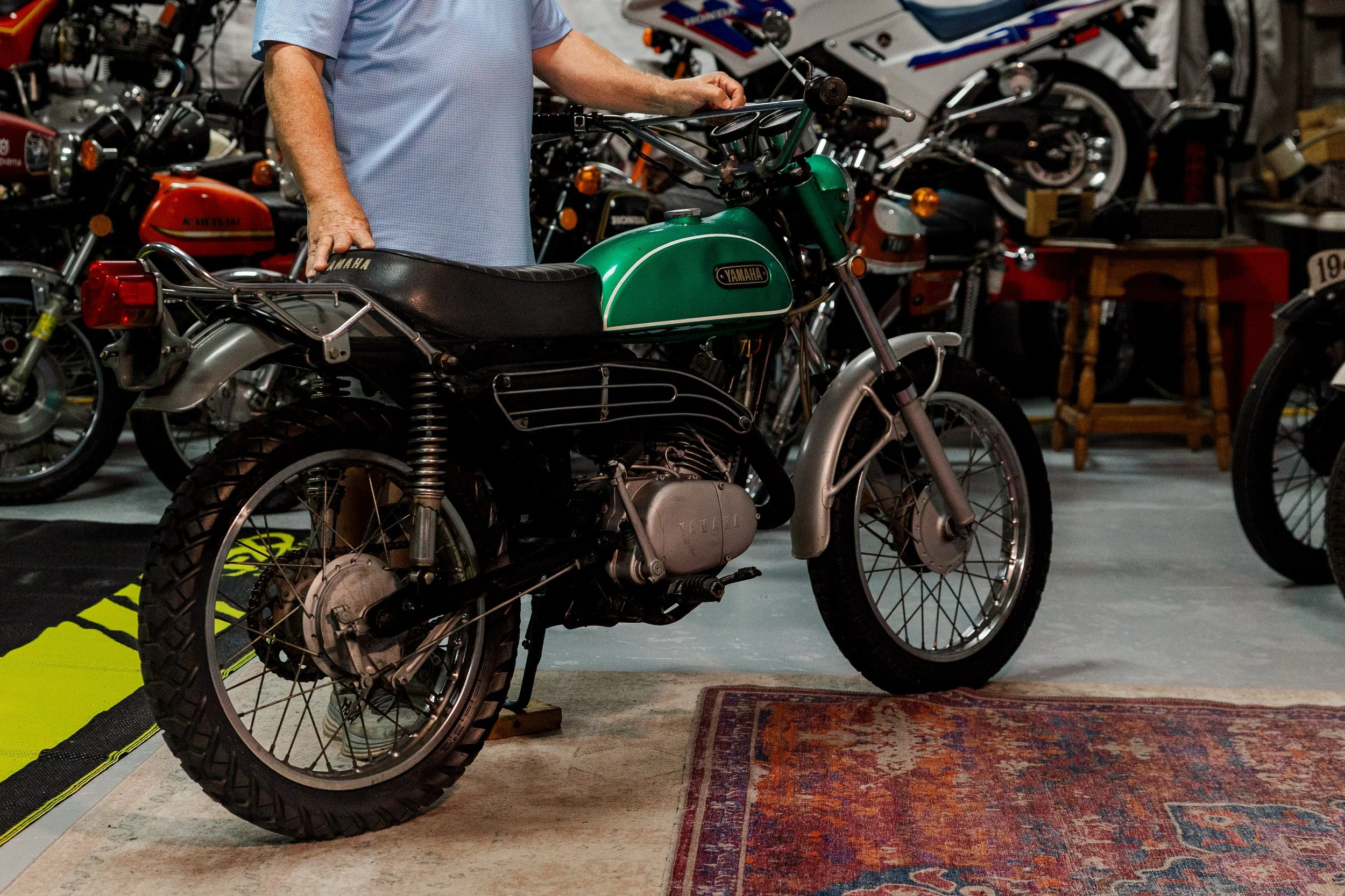 Person inspecting a vintage green Yamaha motorcycle inside a collection or showroom, with multiple other motorcycles visible in the background. Vintage motorcycle collection on display at Birmingham Vintage Cycle in Birmingham Alabama