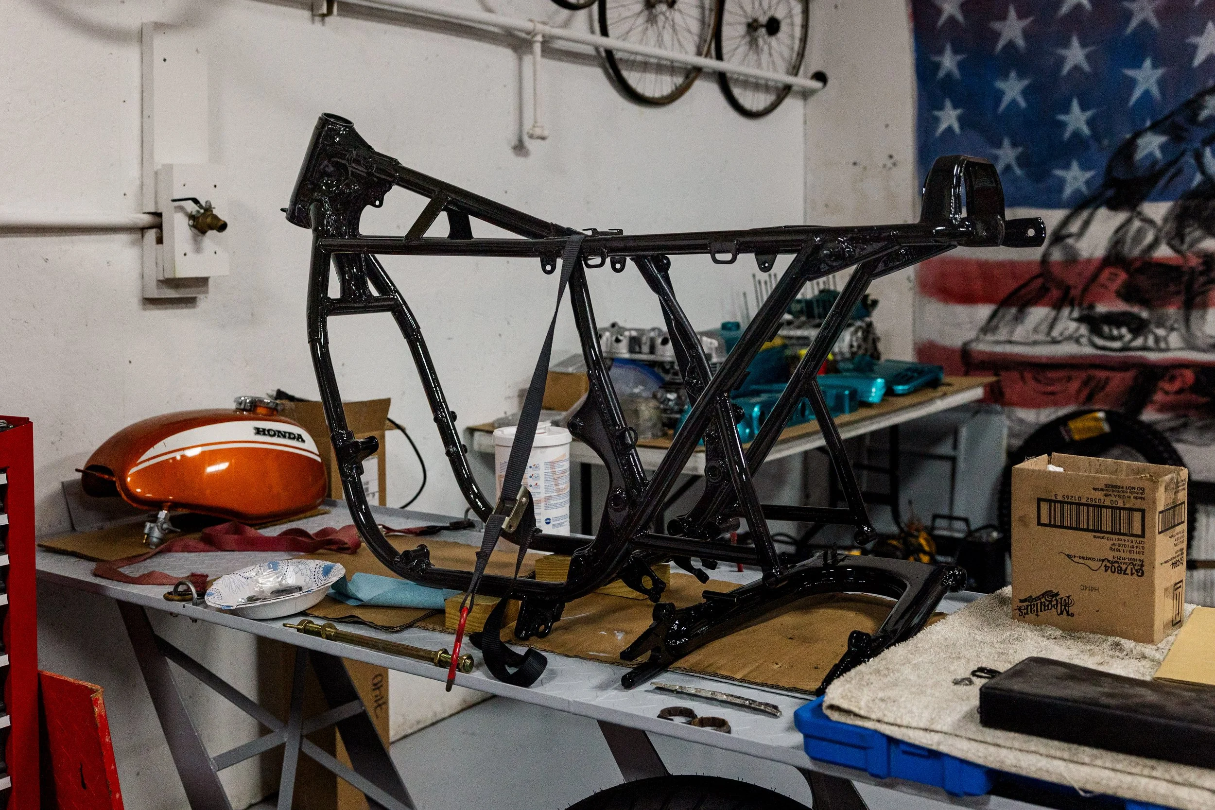 A workbench in a garage with a black painted bicycle frame, an orange Honda motorcycle fuel tank, and various tools and parts scattered on the surface. There's a bicycle wheel mounted on the wall and an American flag hanging in the background.