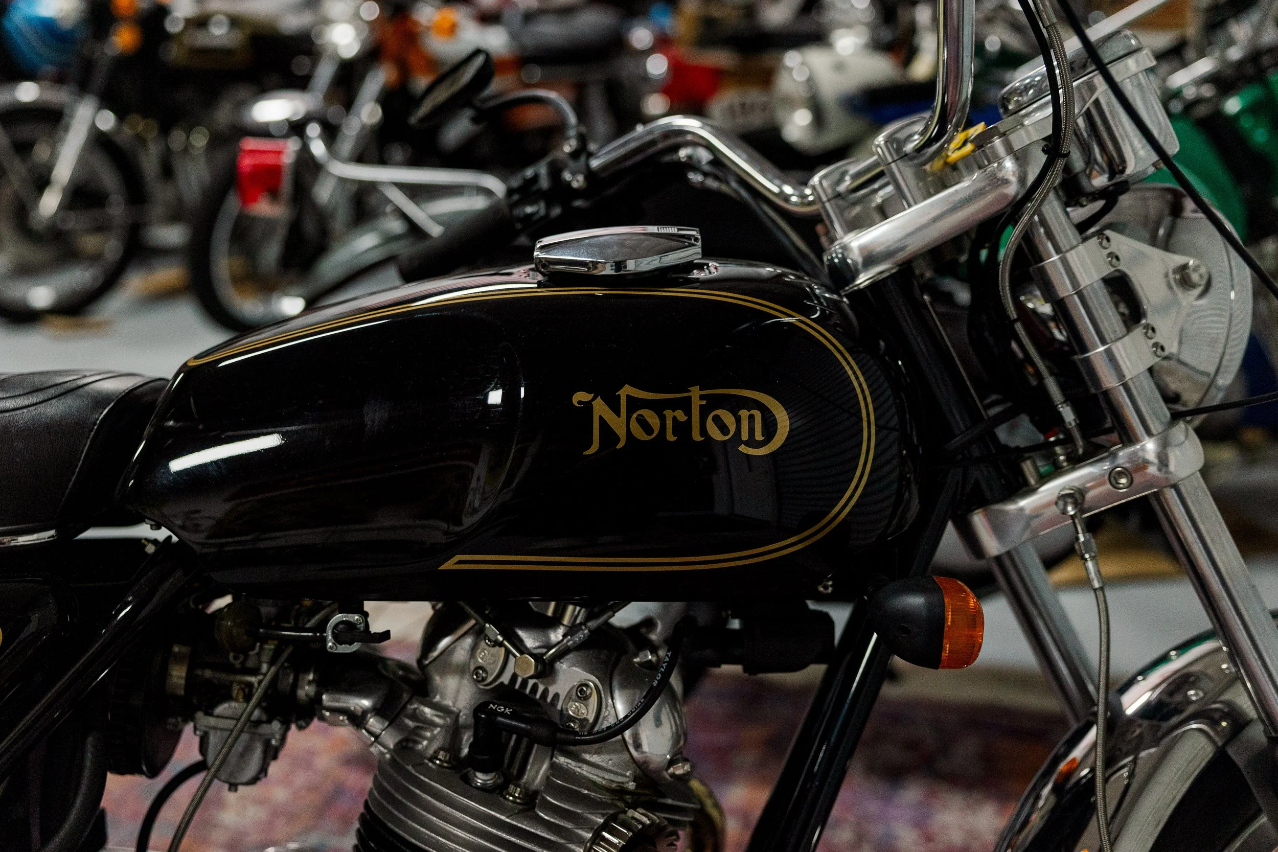 Close-up of a black Norton motorcycle's gas tank, showing chrome details and part of the engine, with other motorcycles blurred in the background. Vintage motorcycle detail at Birmingham Vintage Cycle, a vintage motorcycle restoration shop in