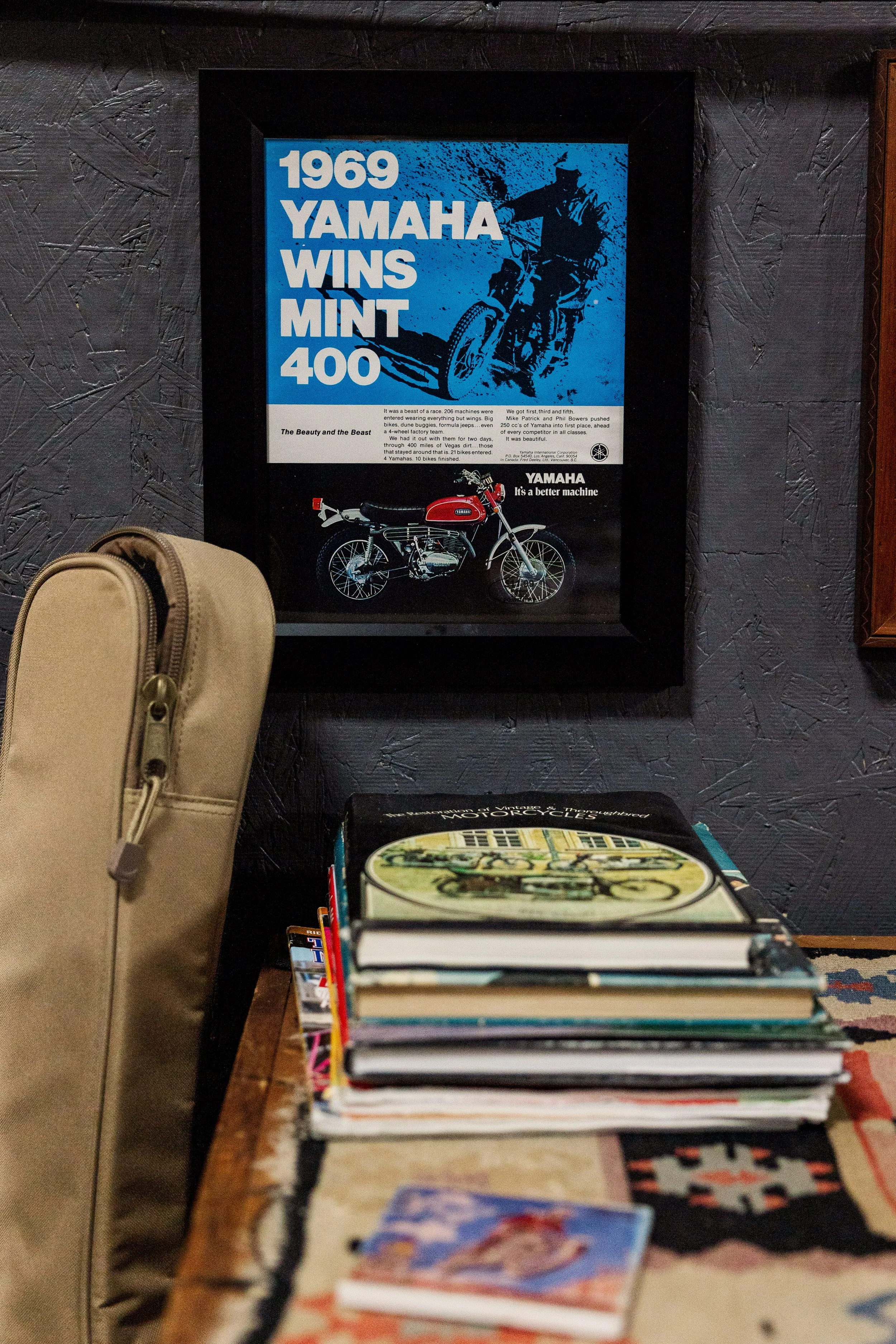 A framed poster on the wall displays a blue and black illustration of a man riding a vintage motorcycle and text celebrating Yamaha's 1969 race wins with models 400 and Mint. In front of the poster, there is a row of children's books stacked on a woo